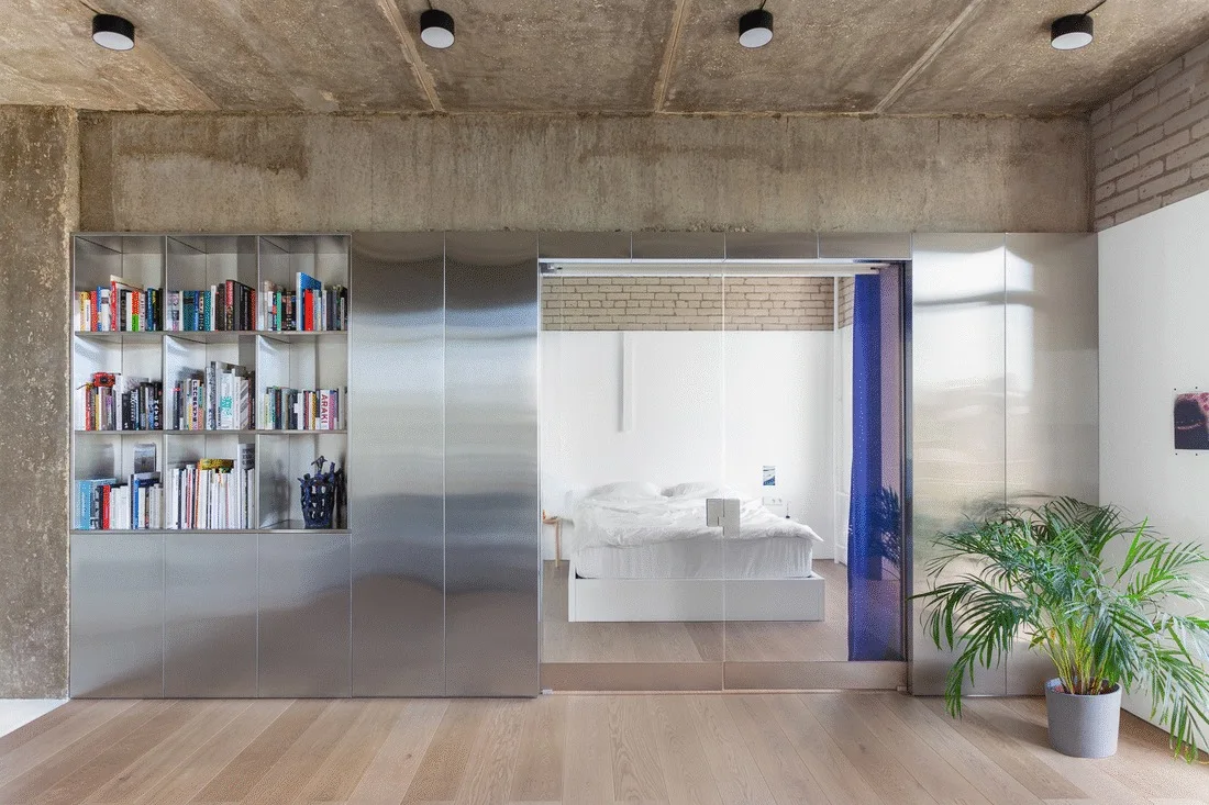 A brushed metal storage unit with an open bookshelf and a glass partition door that opens into a minimal white bedroom under a raw concrete ceiling.