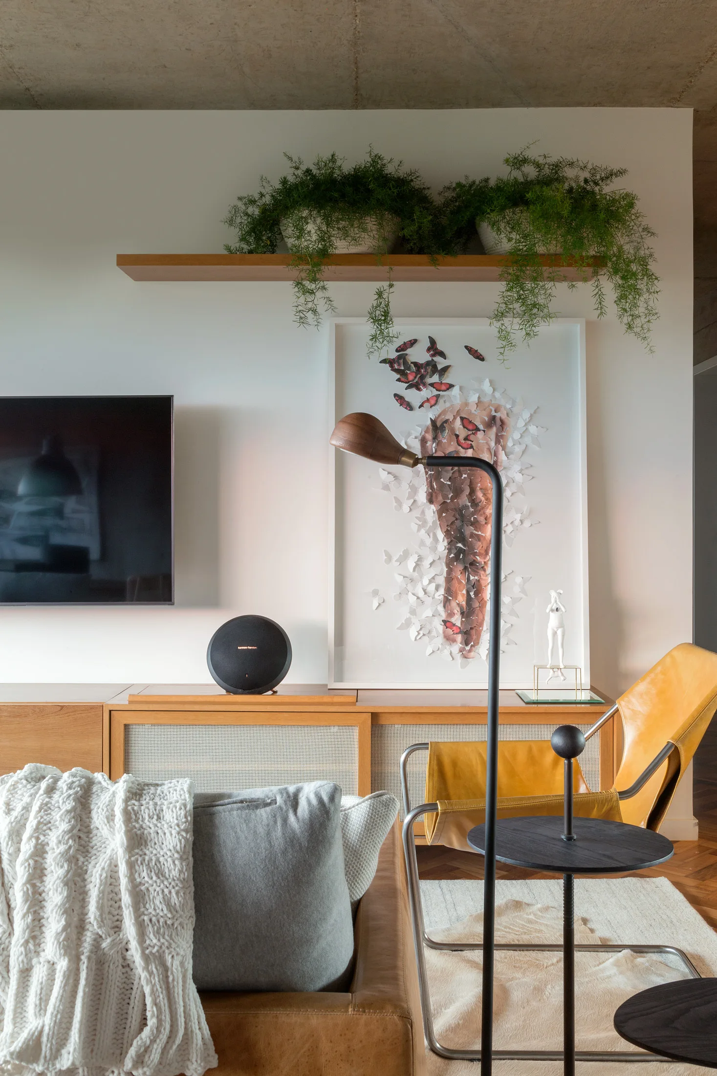 A modern living room featuring a leather sofa, a wood and cane media console, a yellow leather sling chair, and cascading plants on a shelf.