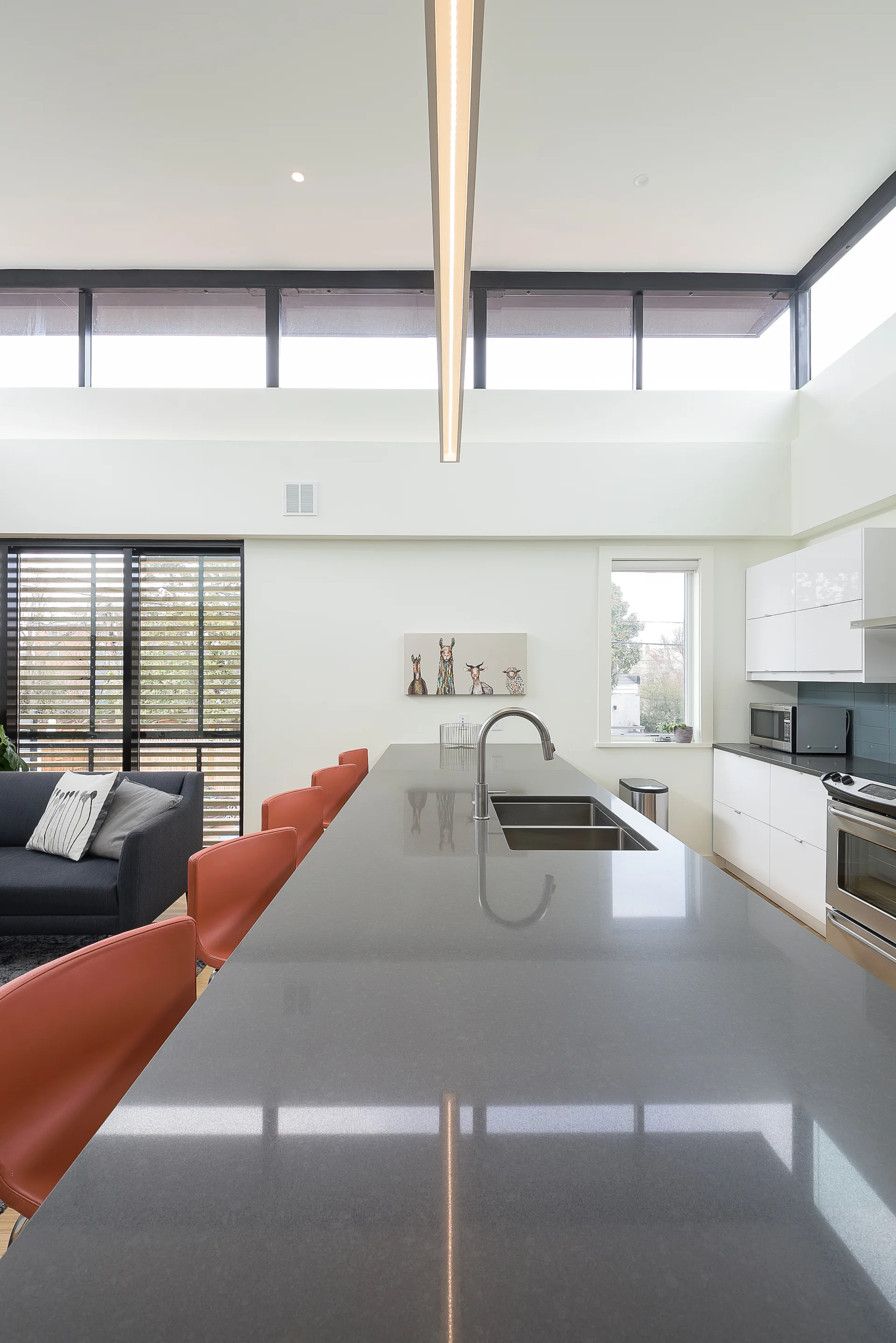 A modern kitchen featuring a long gray stone island with a double sink, orange bar stools, glossy white cabinets, and high clerestory windows.