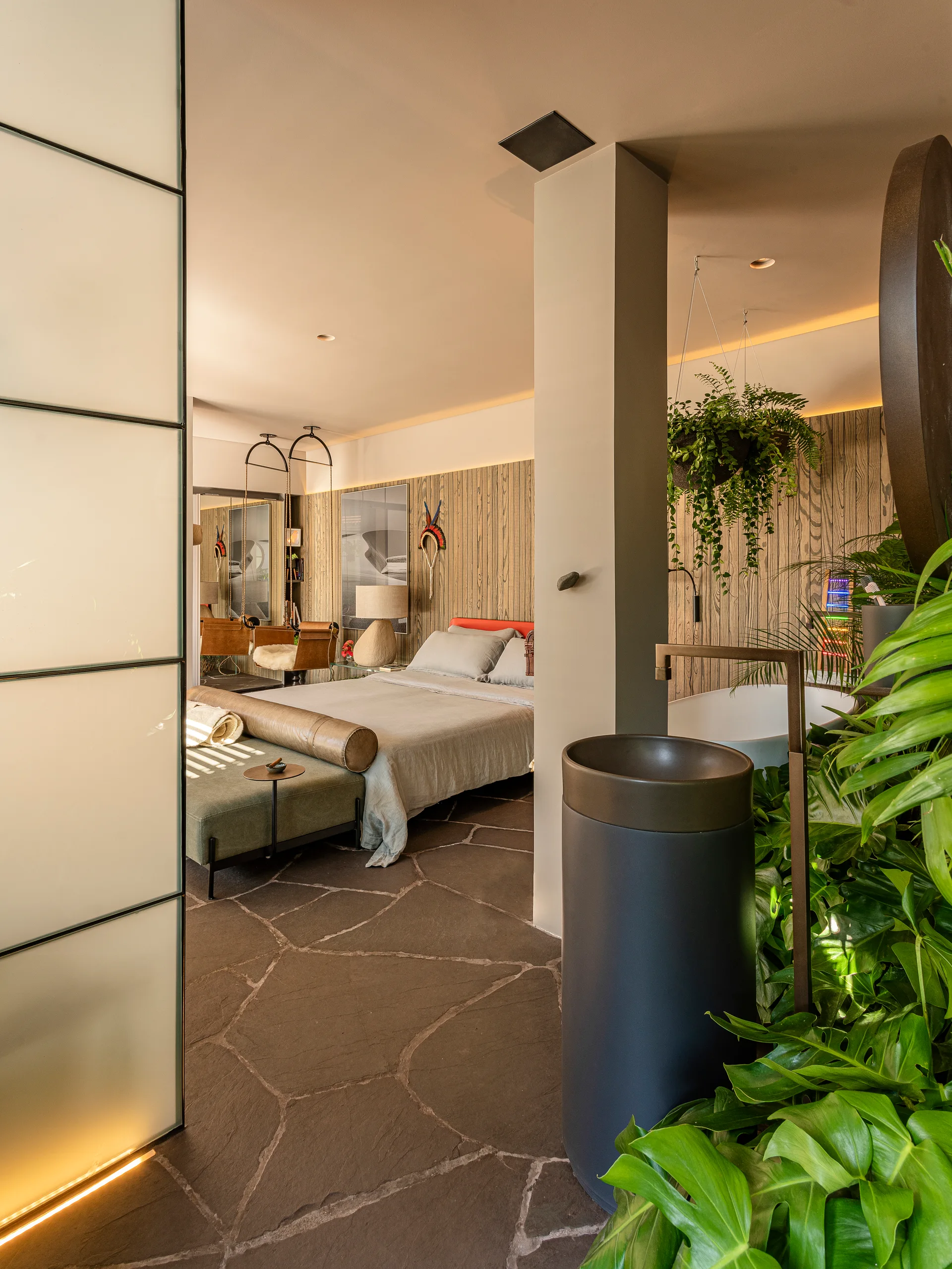 An open-plan bedroom features flagstone flooring, wood-paneled walls, a green bed bench, and an integrated bathroom sink surrounded by plants.