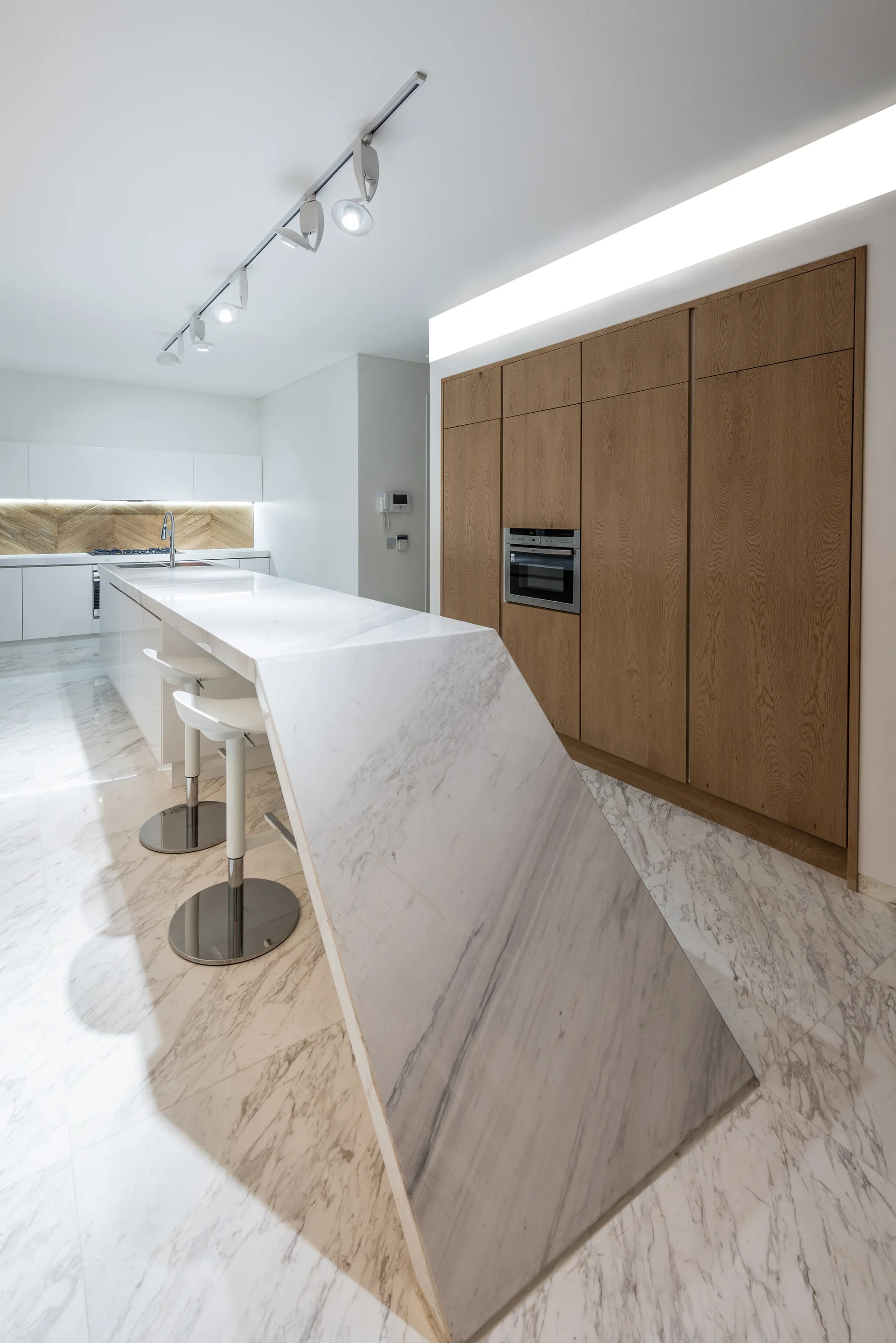 A modern kitchen with a white marble waterfall island, matching marble floors, white bar stools, and warm wood wall cabinets.