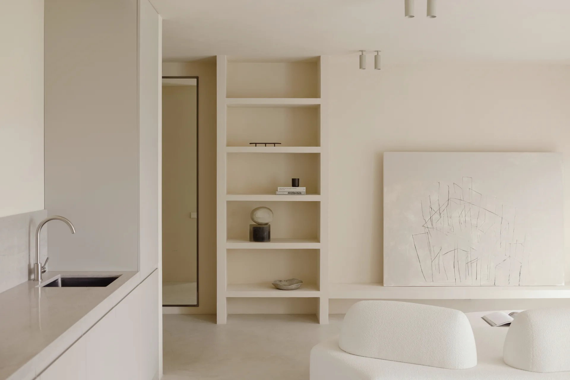A minimalist beige living room featuring an integrated wall shelving unit, a large leaning painting, a curved boucle sofa, and a kitchen counter.