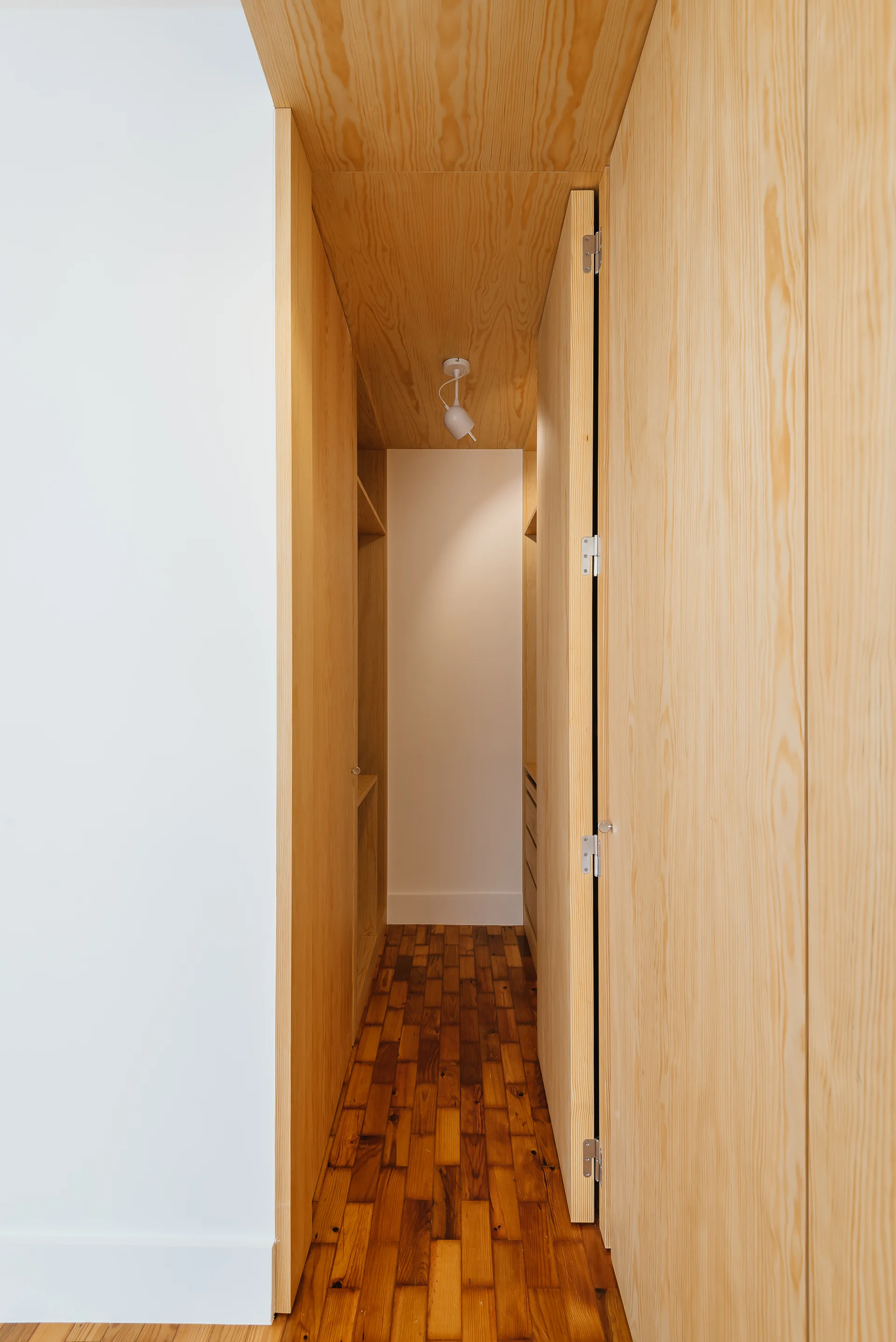 A narrow walk-in closet features light wood clad walls and ceiling, custom plywood cabinetry, and dark wood block parquet flooring.