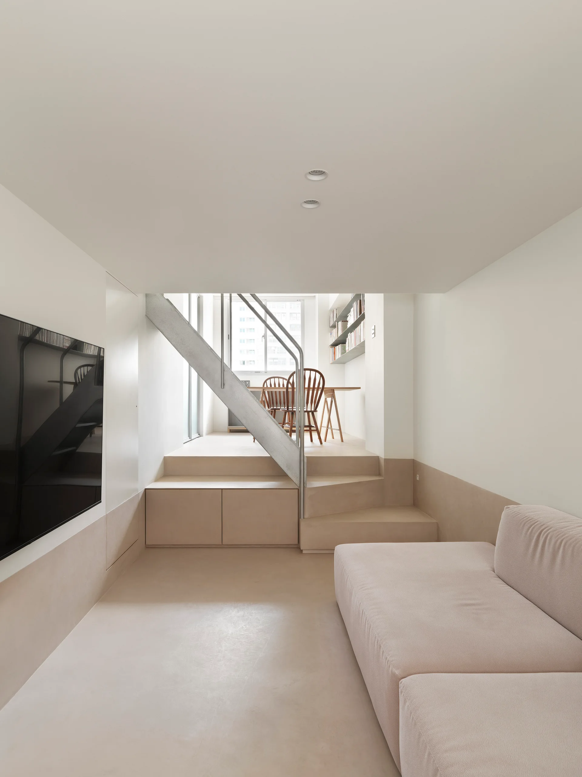 A minimalist split-level living room features a beige low-profile sofa, a flat-screen TV, and galvanized metal stairs leading to an elevated dining area.