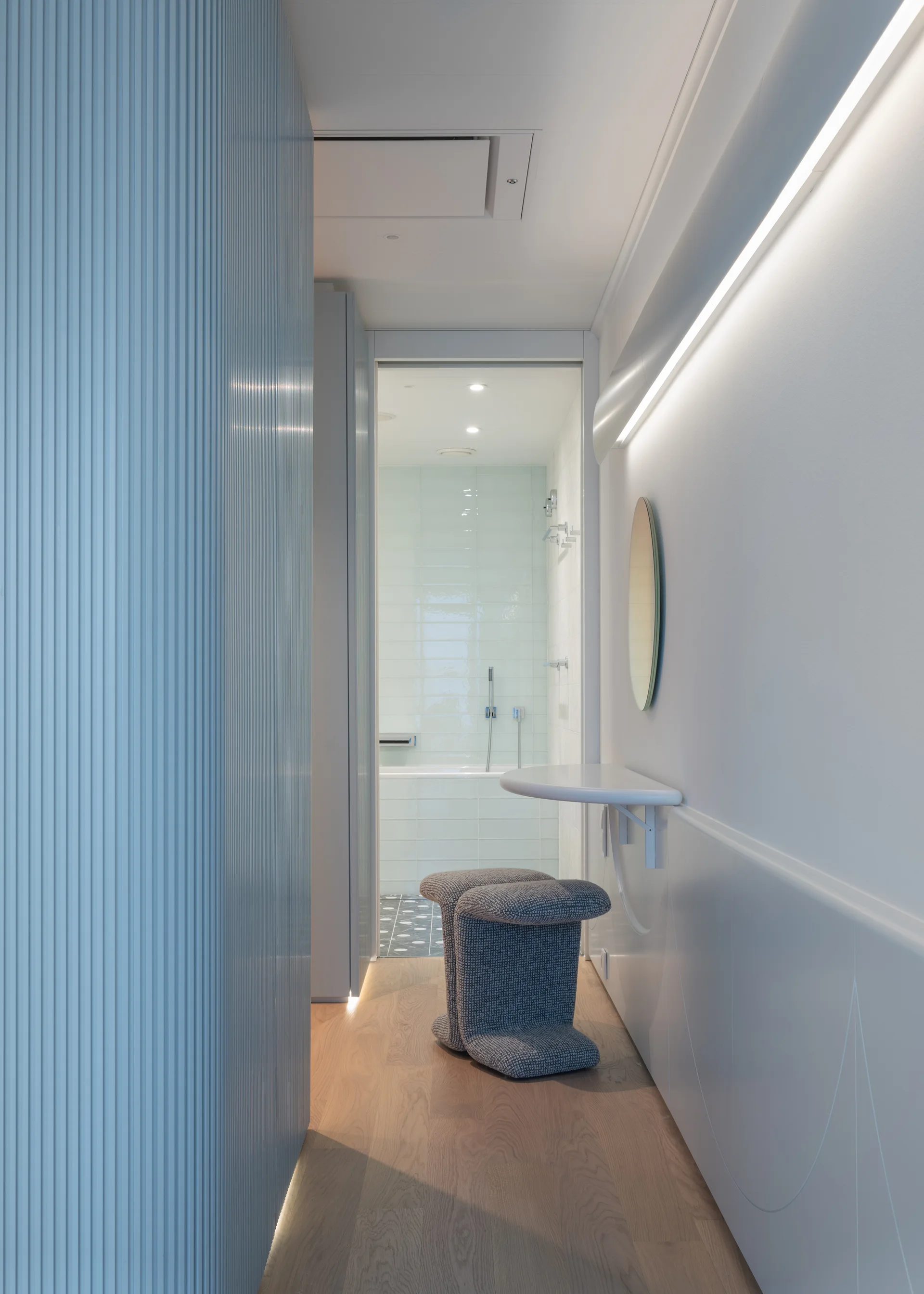 A modern hallway features a light blue fluted wall, a floating white semi-circular vanity table with a grey textured stool, leading to a tiled bathroom.