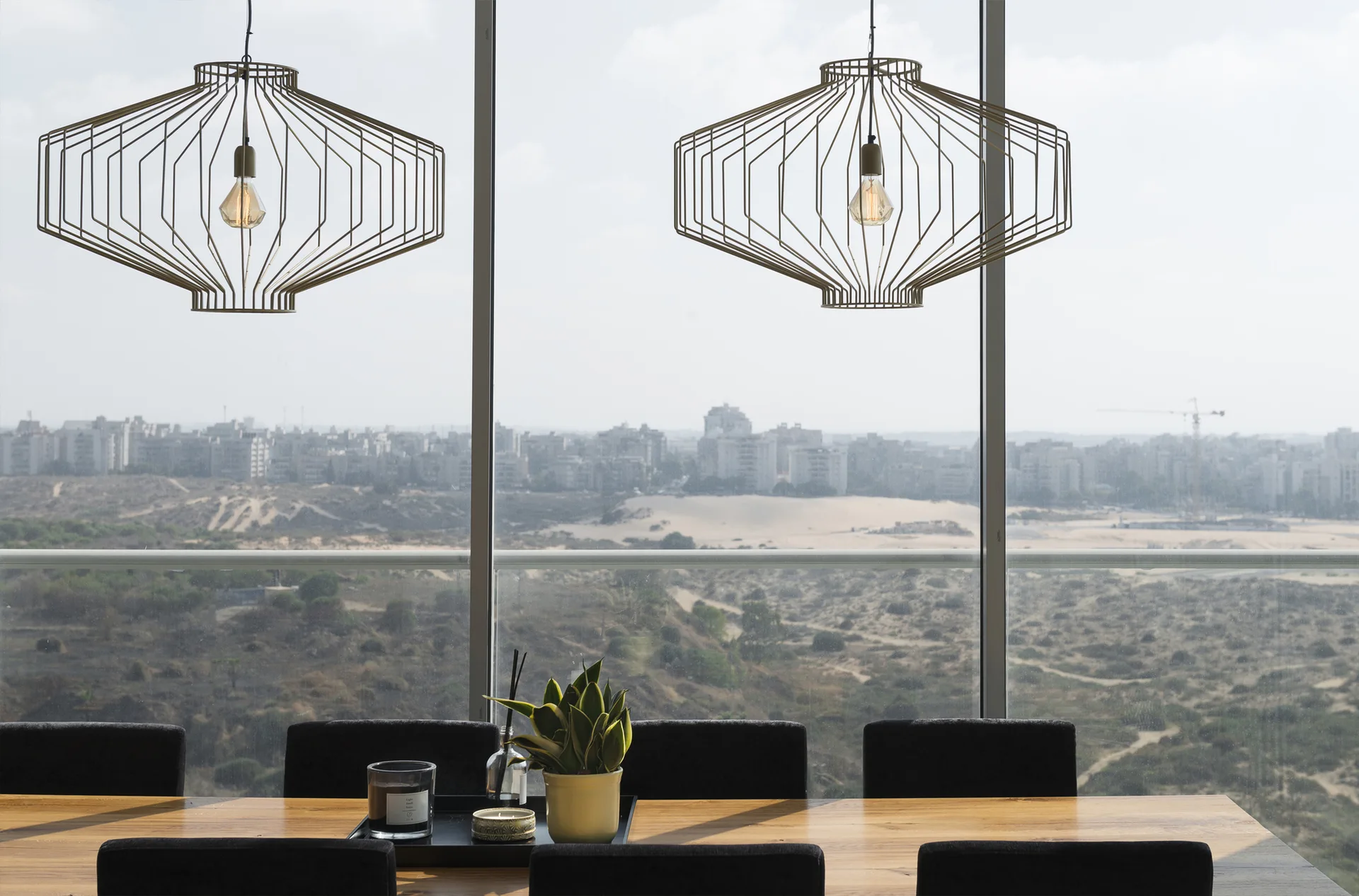 Two geometric wire pendant lights hang above a wooden dining table with dark chairs in front of large windows showing an expansive landscape.