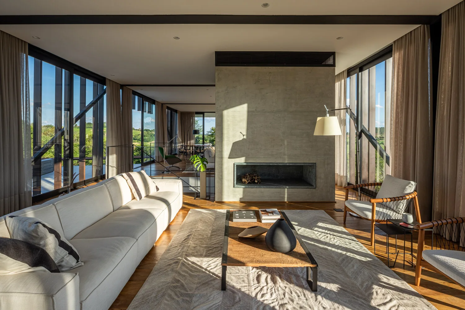 A modern living room featuring a cream sofa, a low wood coffee table, and a central concrete fireplace partition between large glass walls.