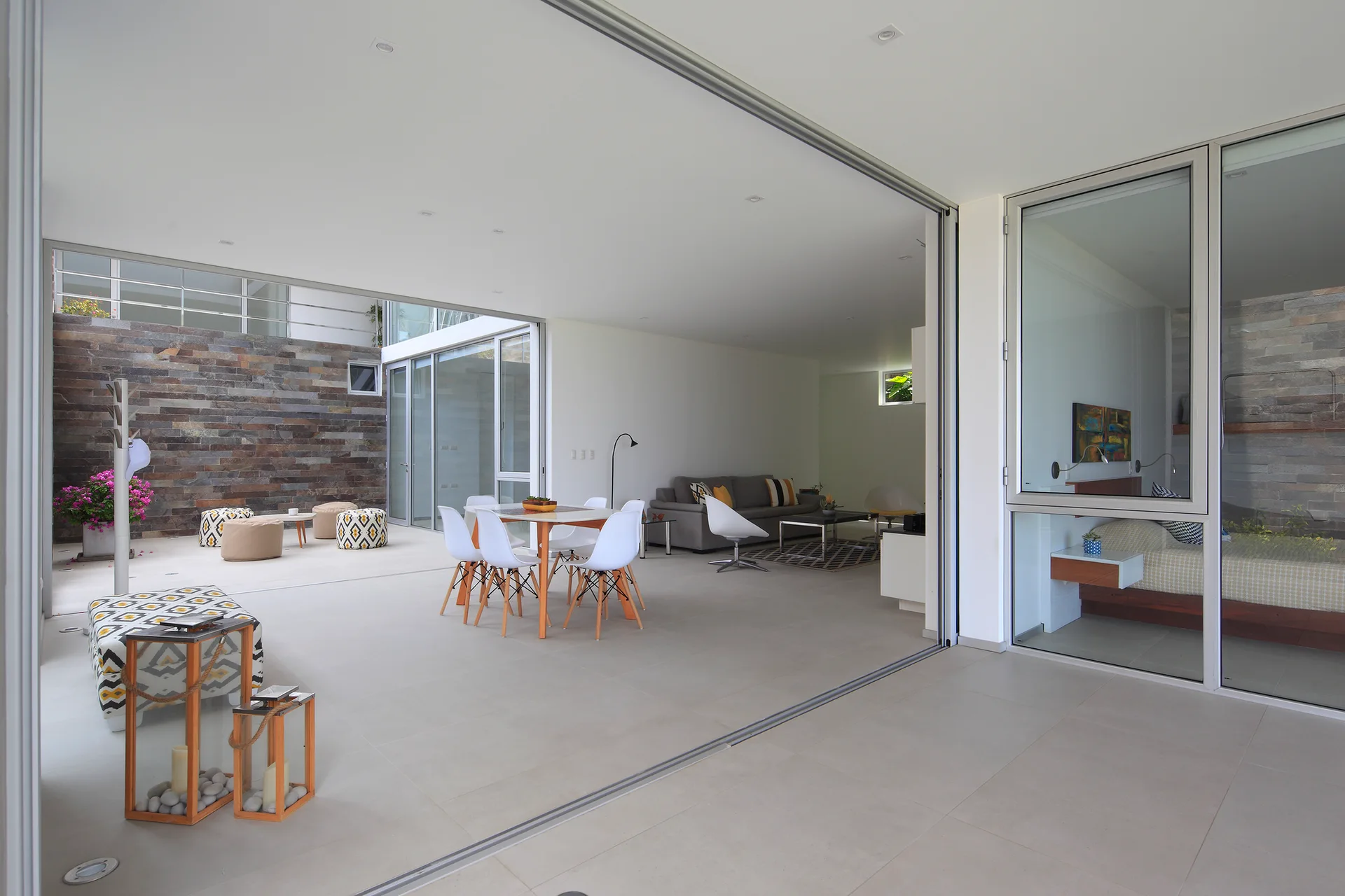 A modern open-concept home features white dining chairs around a table next to large retractable glass doors opening to a stone-walled courtyard.
