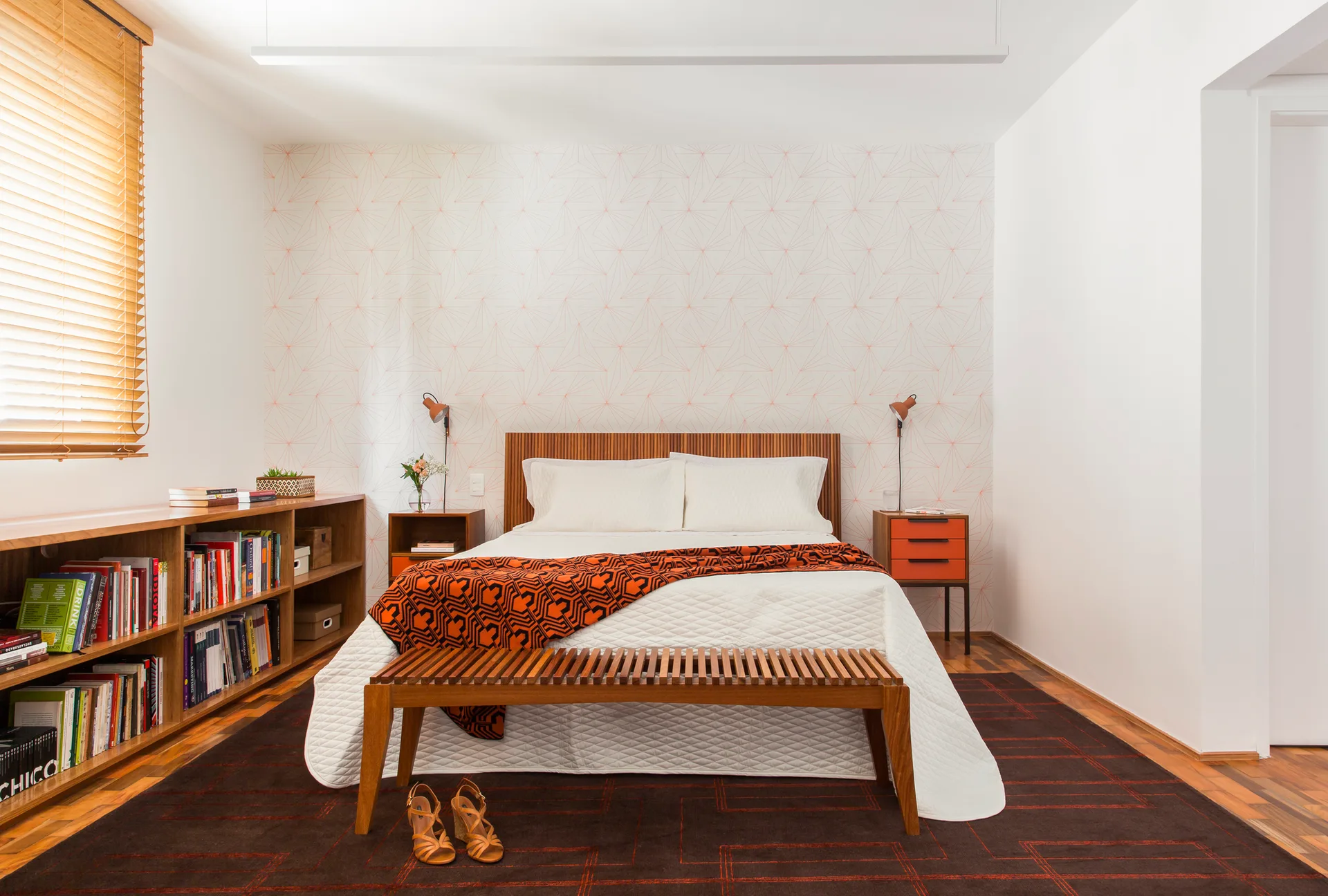 A mid-century modern bedroom features a wooden bed with a slatted headboard and bench, orange accents, geometric wallpaper, and a wooden bookcase.