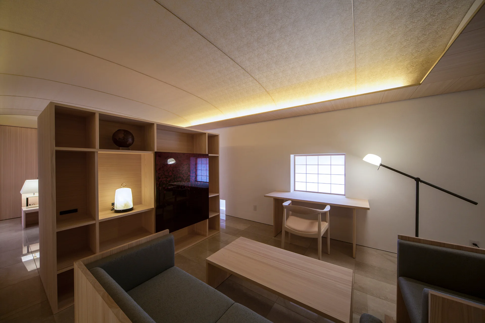 A minimalist living room featuring a curved ceiling with cove lighting, a wood bookshelf divider, and a desk under a grid window.