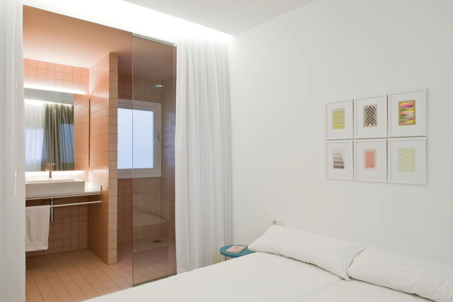A minimalist bedroom with a white bed sits next to an open-plan bathroom tiled in pastel pink squares with a glass partition.
