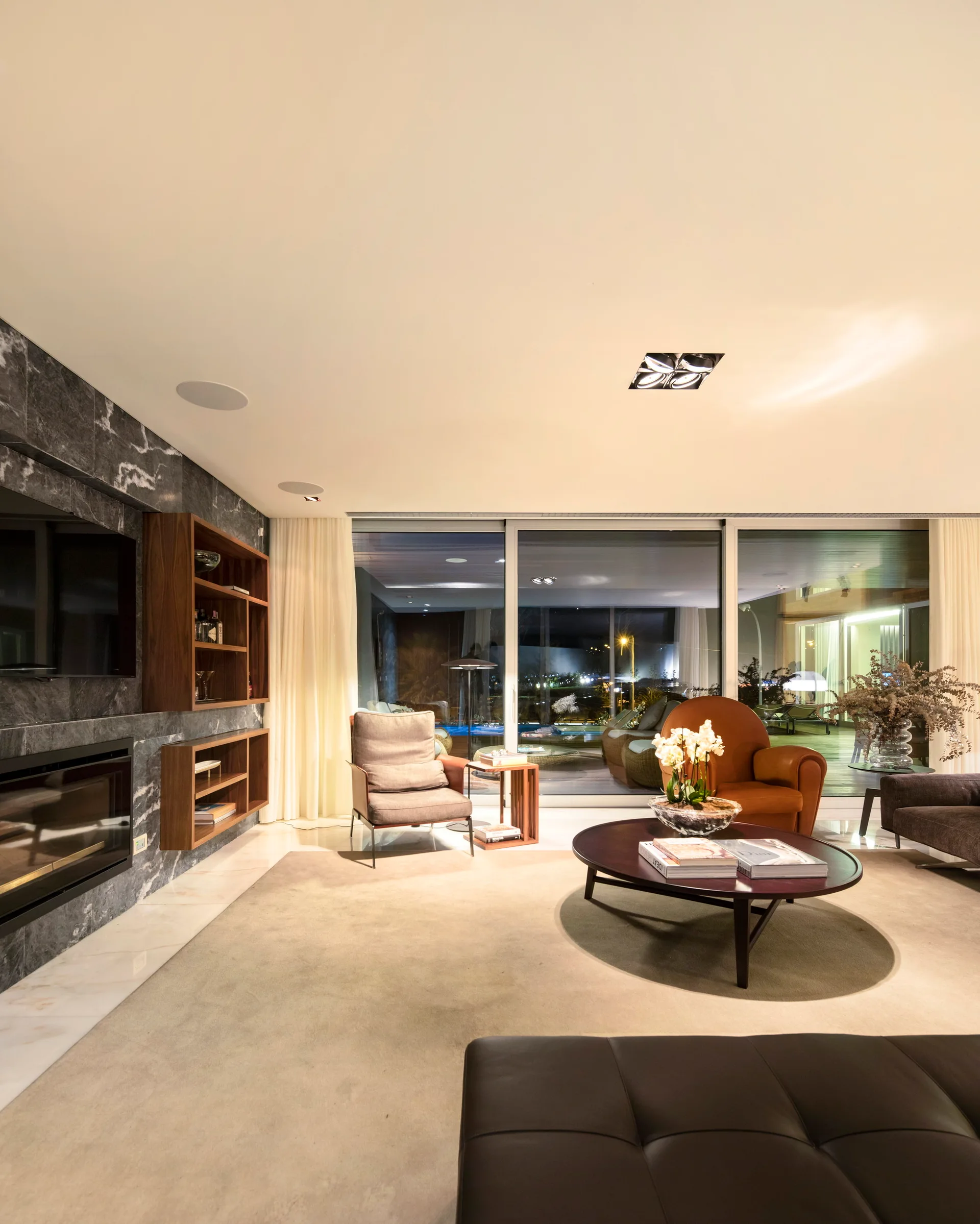 A modern living room with a dark marble fireplace, floating wood shelves, a circular wood coffee table, and large glass sliding doors opening to a patio.