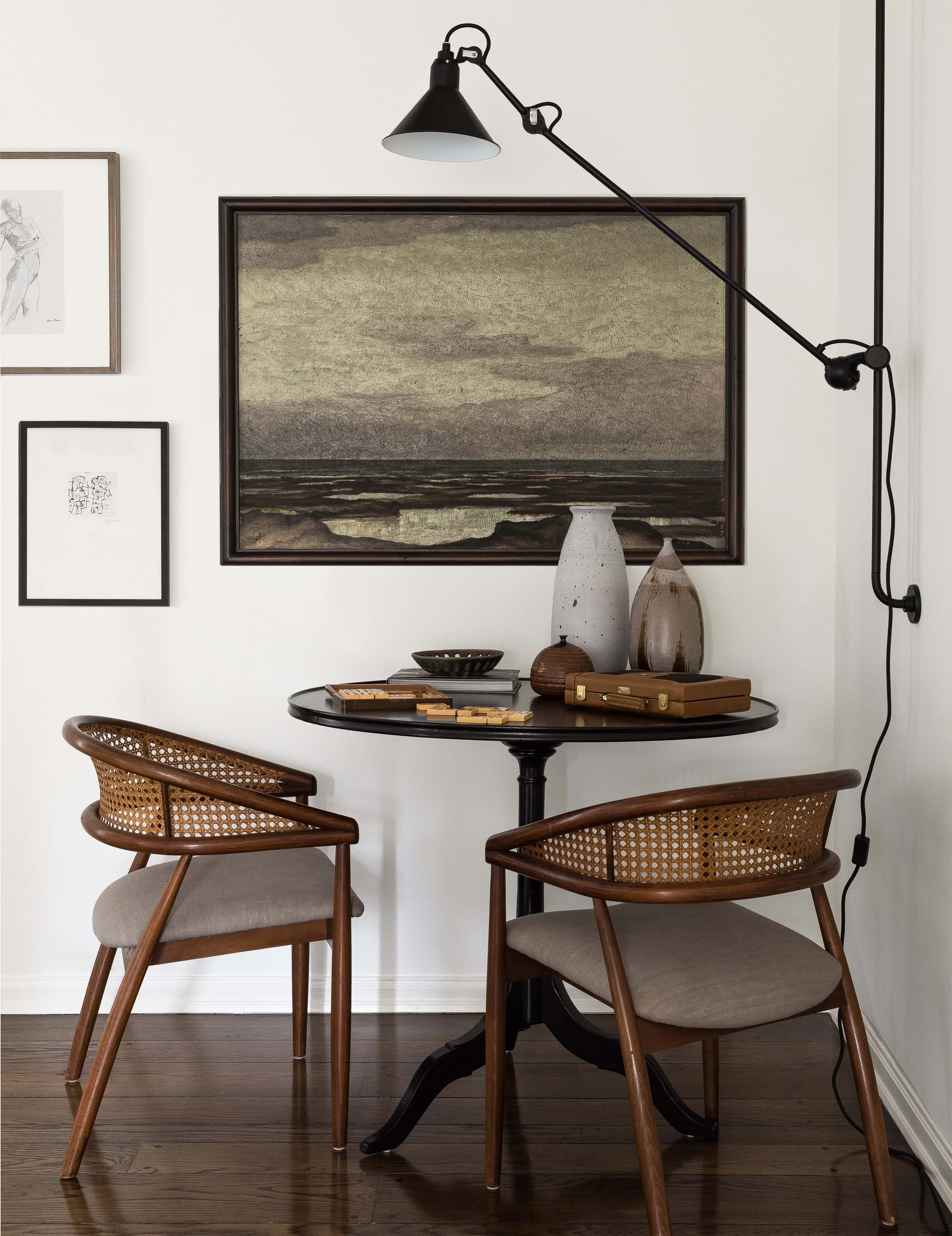Two wood and cane chairs face a black pedestal table in a room corner below framed artwork and a black swing-arm wall lamp.