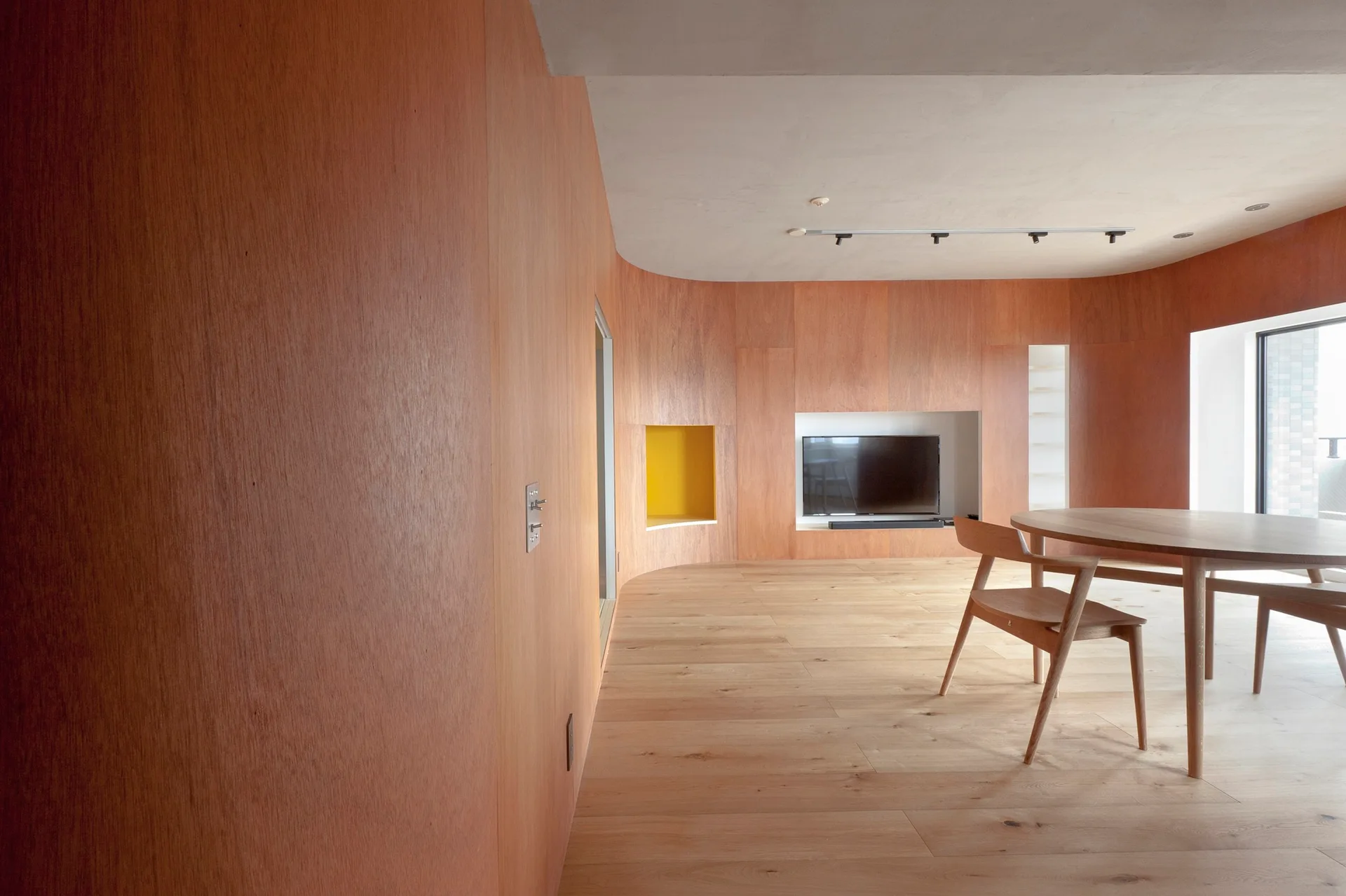 A minimalist dining room with curved plywood walls, a round wooden table, chairs, and recessed white and yellow niches.