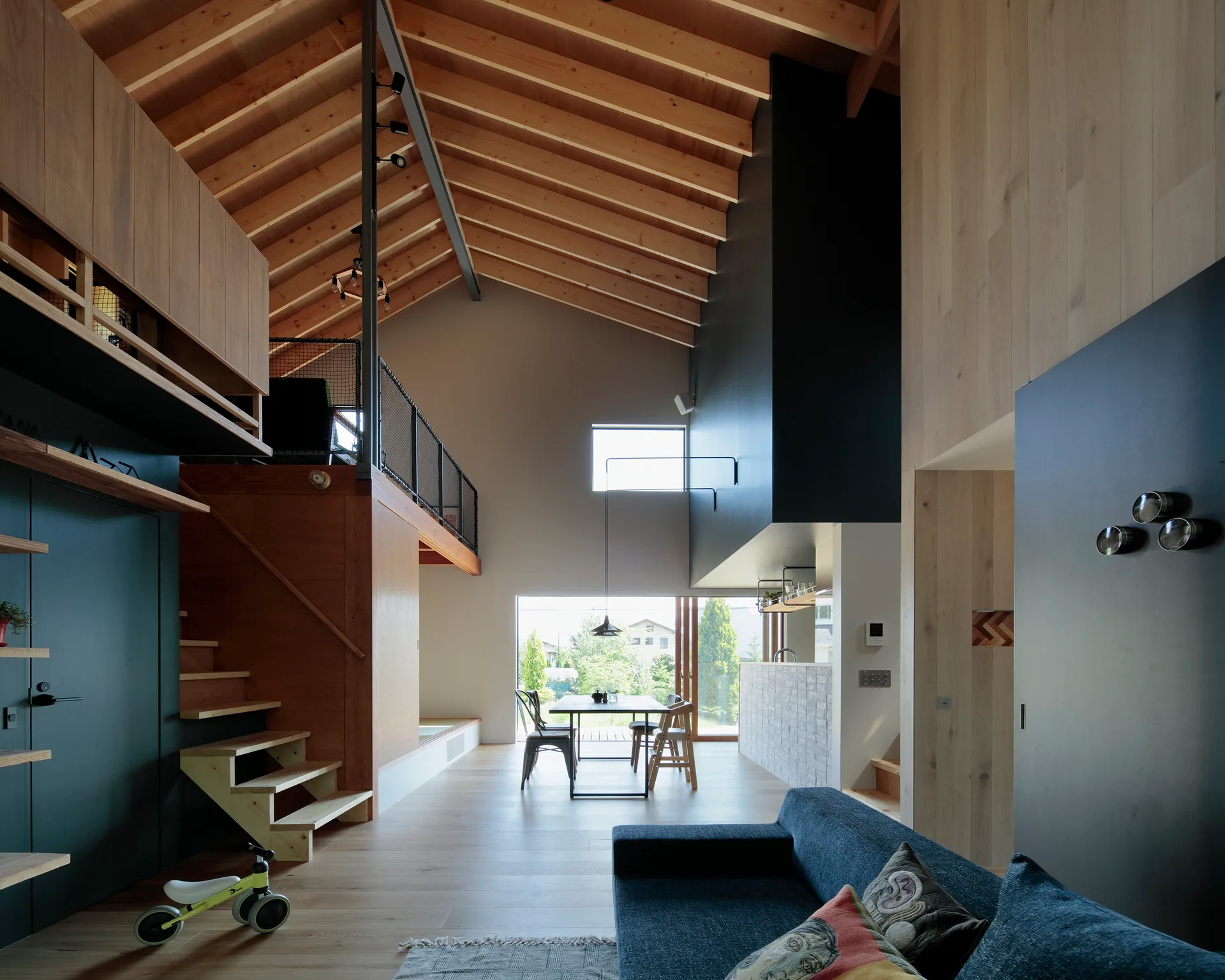 A modern open-plan living room featuring a vaulted wood ceiling, a mezzanine loft, an open staircase, and a dining area in the background.