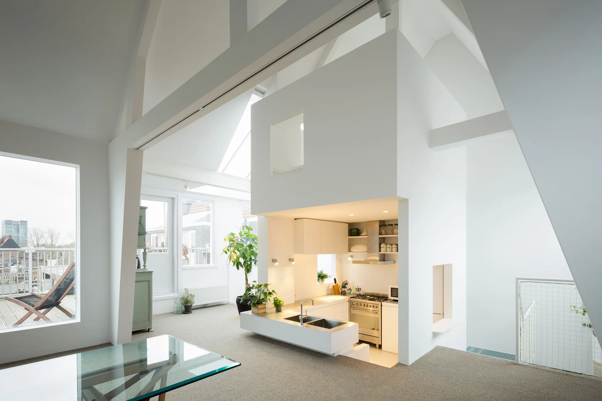 A minimalist open-plan loft features a central white kitchen block beneath a vaulted ceiling with skylights and tall windows.