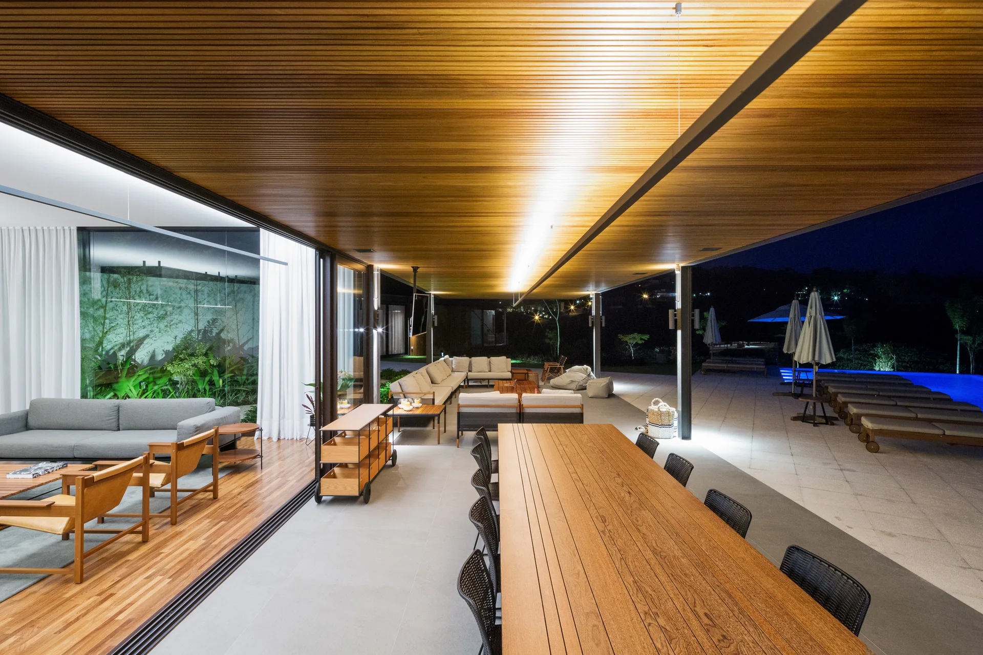 A spacious covered terrace features a slatted wood ceiling, a long dining table, and lounge furniture next to a swimming pool.