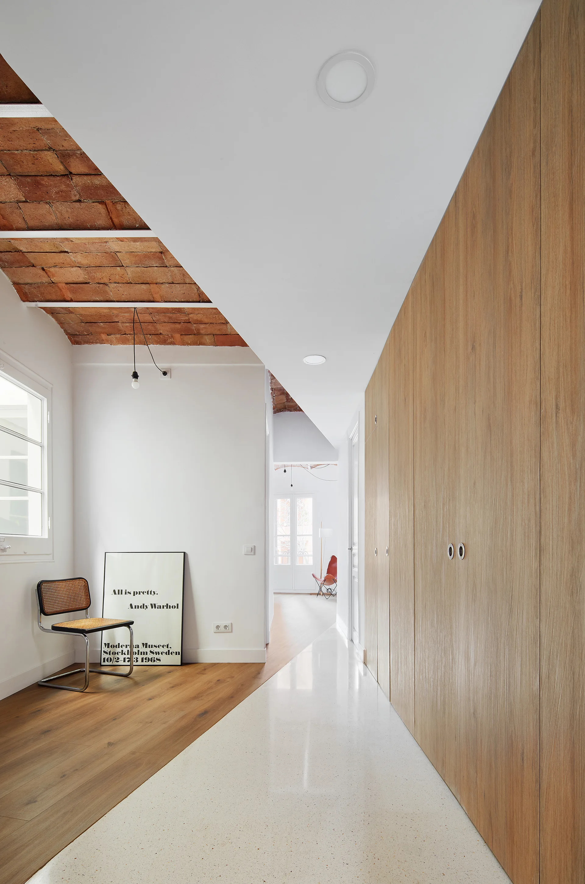 A modern hallway features a vaulted brick ceiling, wood paneled closets, a cane chair, and a floor divided diagonally into wood and speckled white terrazzo.