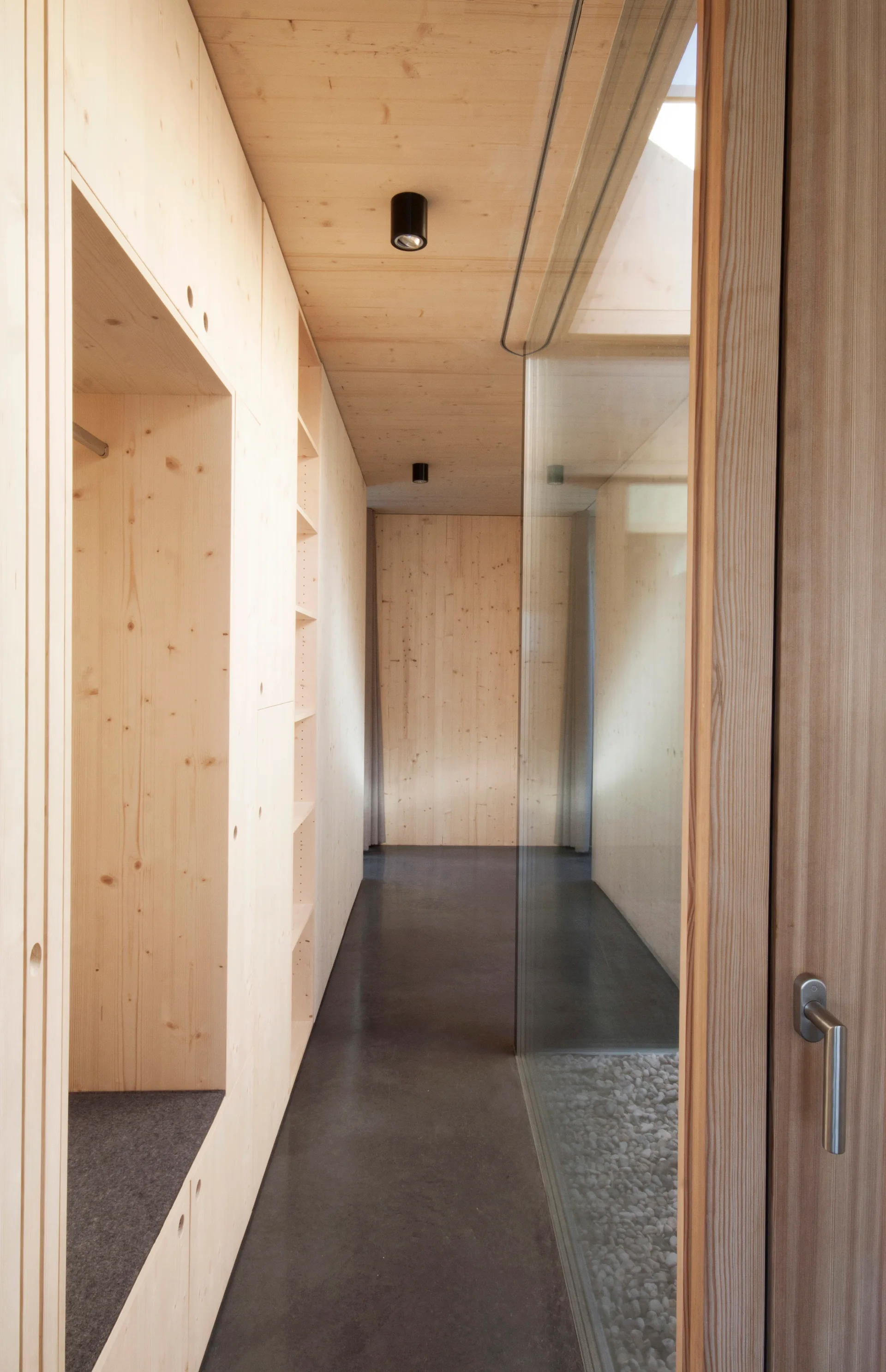 A minimalist hallway featuring light wood paneled walls and ceiling, a smooth dark concrete floor, and a glass partition looking into a pebble bed.