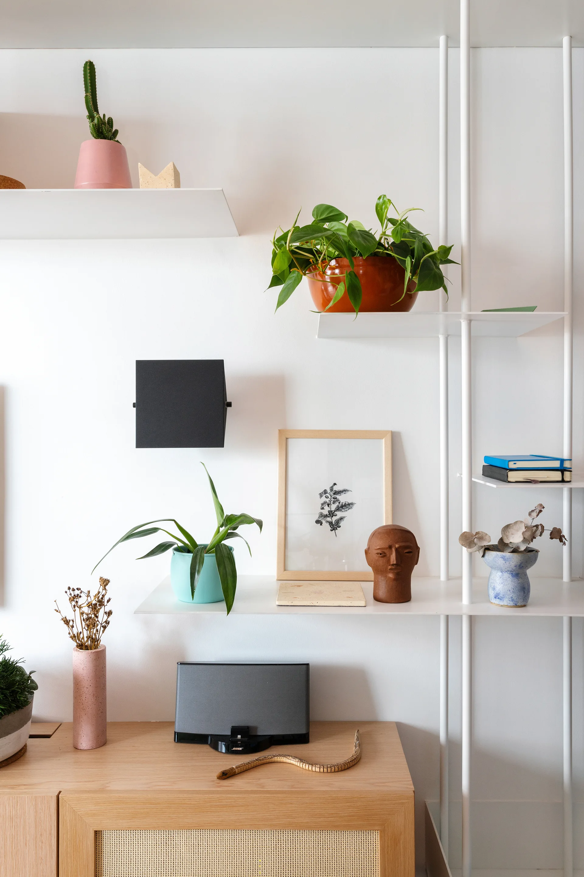 A white metal shelving system displays potted plants and art above a light wood console cabinet with woven cane doors against a plain white wall.