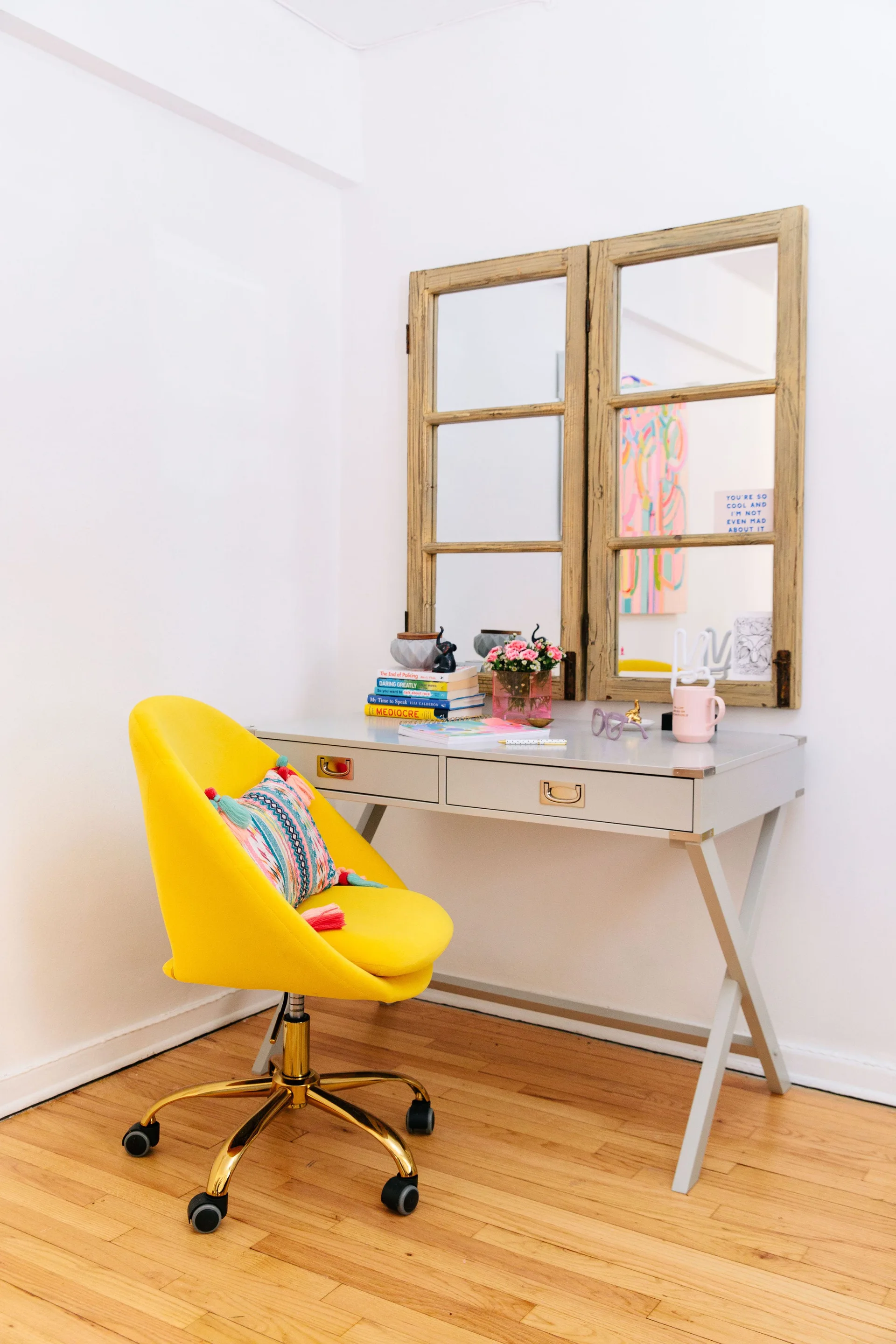 A bright yellow swivel chair sits at a light grey writing desk beneath a rustic wooden window-frame mirror on a white wall.