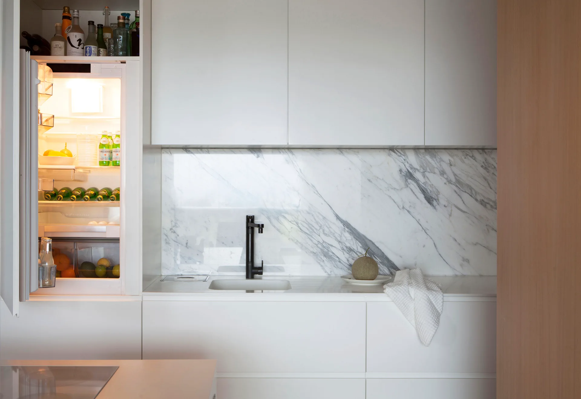 A minimalist white kitchen featuring flat-panel cabinets, a veined marble backsplash, a black faucet, and an open integrated refrigerator.