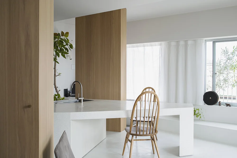 A minimalist kitchen features a geometric white island with a sink, two wood spindle-back chairs, and vertical wood wall cladding.