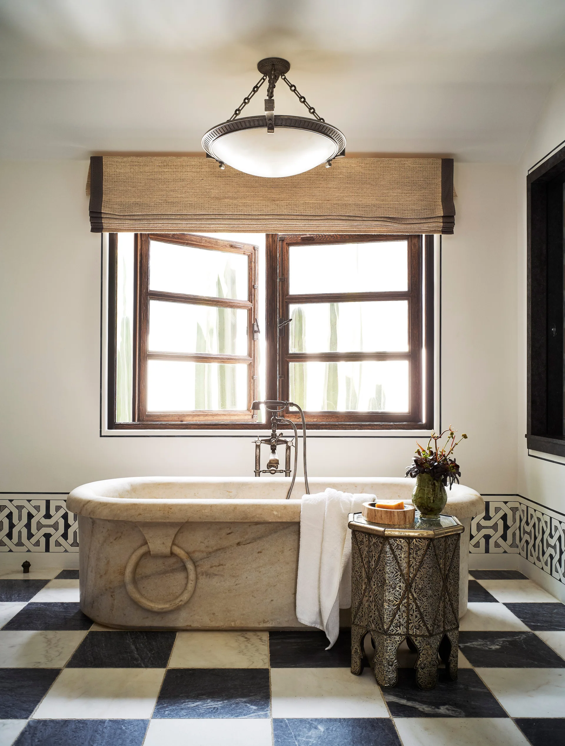 A carved stone bathtub sits in front of a wooden casement window in a bathroom with a black and white checkerboard marble floor.
