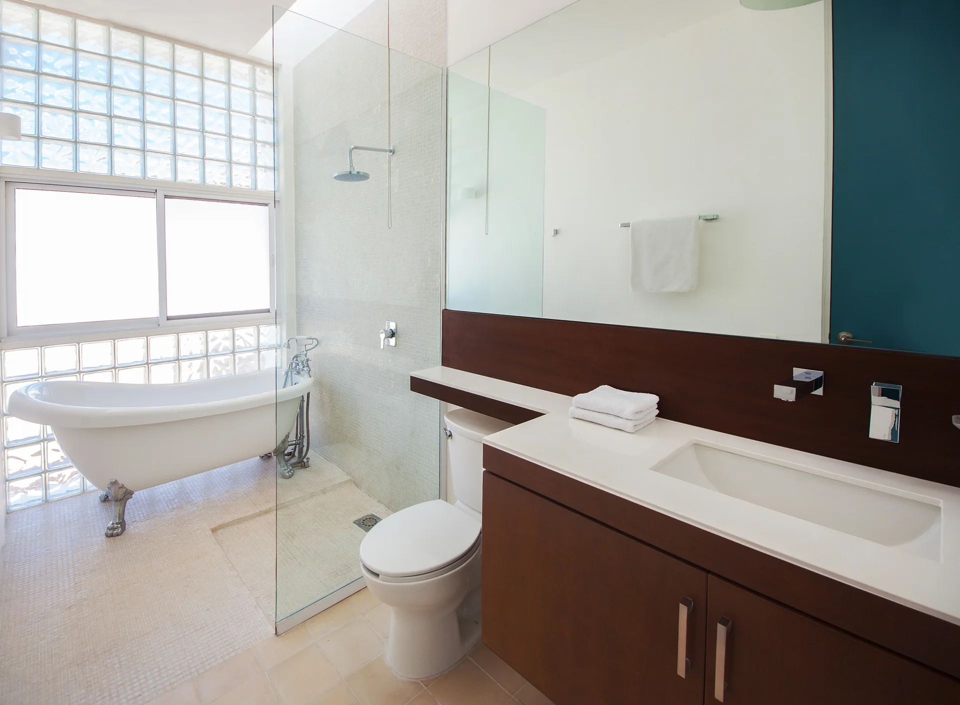 A modern bathroom features a white clawfoot bathtub next to a glass block window, a dark wood vanity, and a clear glass partition.