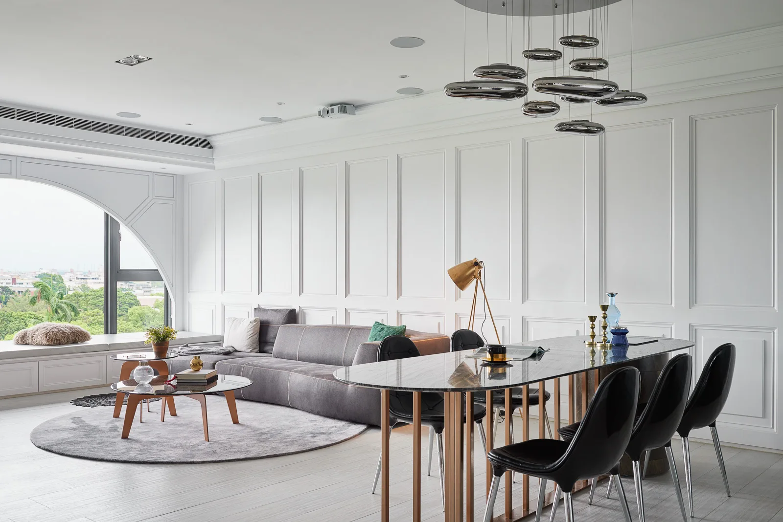 A modern open-plan living space with white wall paneling, a grey sectional sofa, and a marble dining table next to an arched window.
