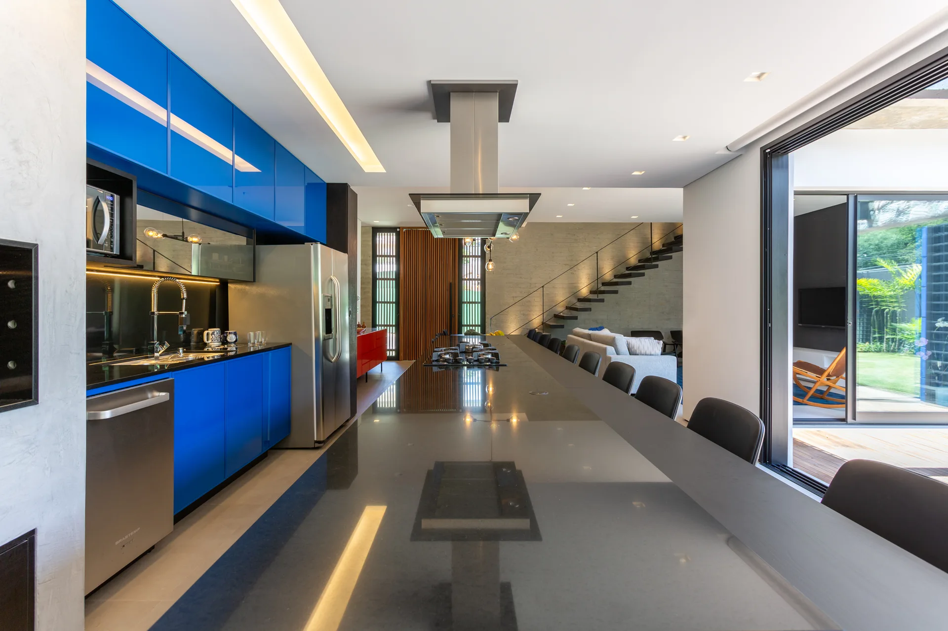 A modern open-plan kitchen featuring high-gloss blue cabinetry, a long dark island with black stools, and a view into a living area.