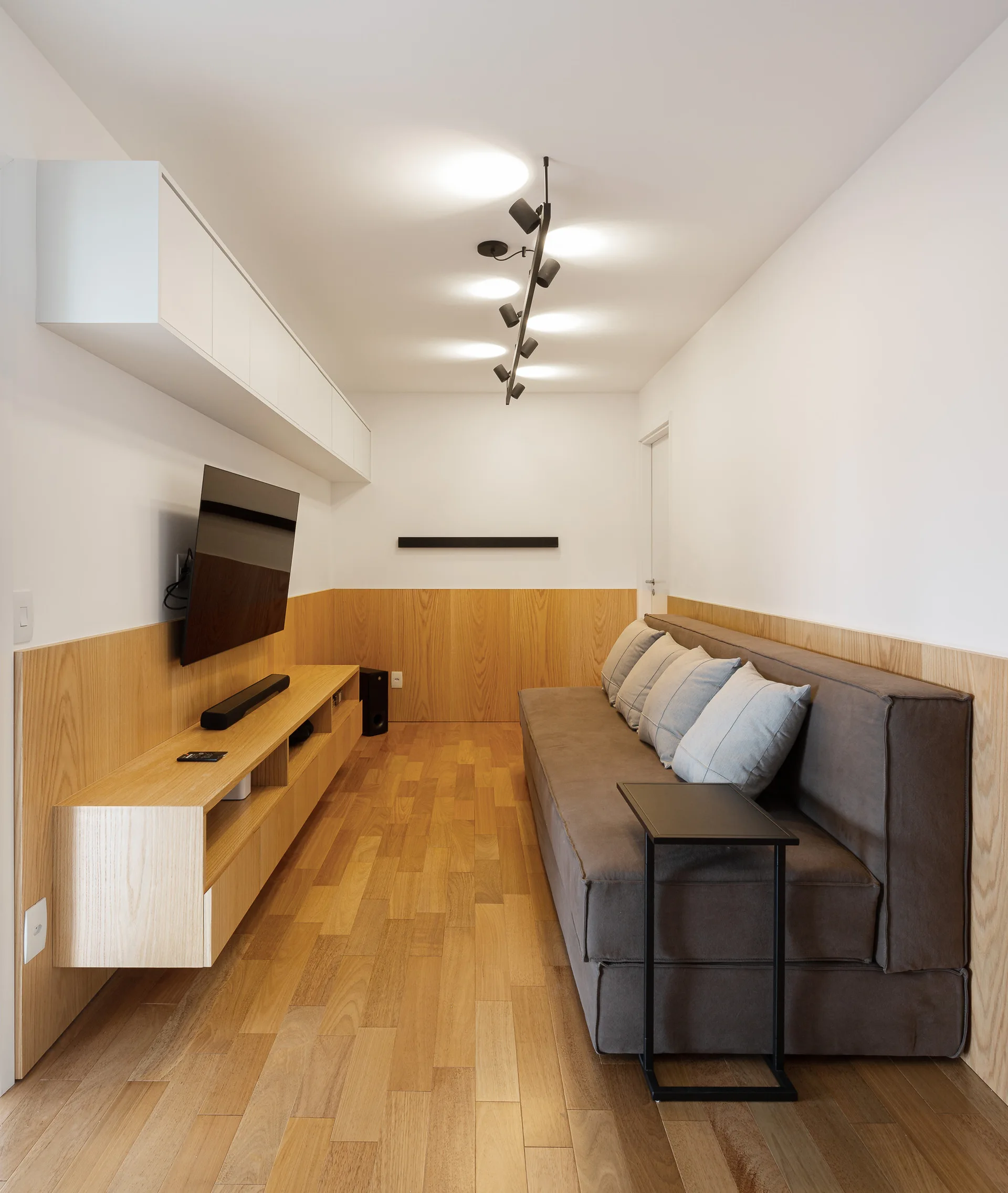A narrow media room features a grey sofa, a floating wood TV console, and warm wood wall paneling running along the lower half of the walls.