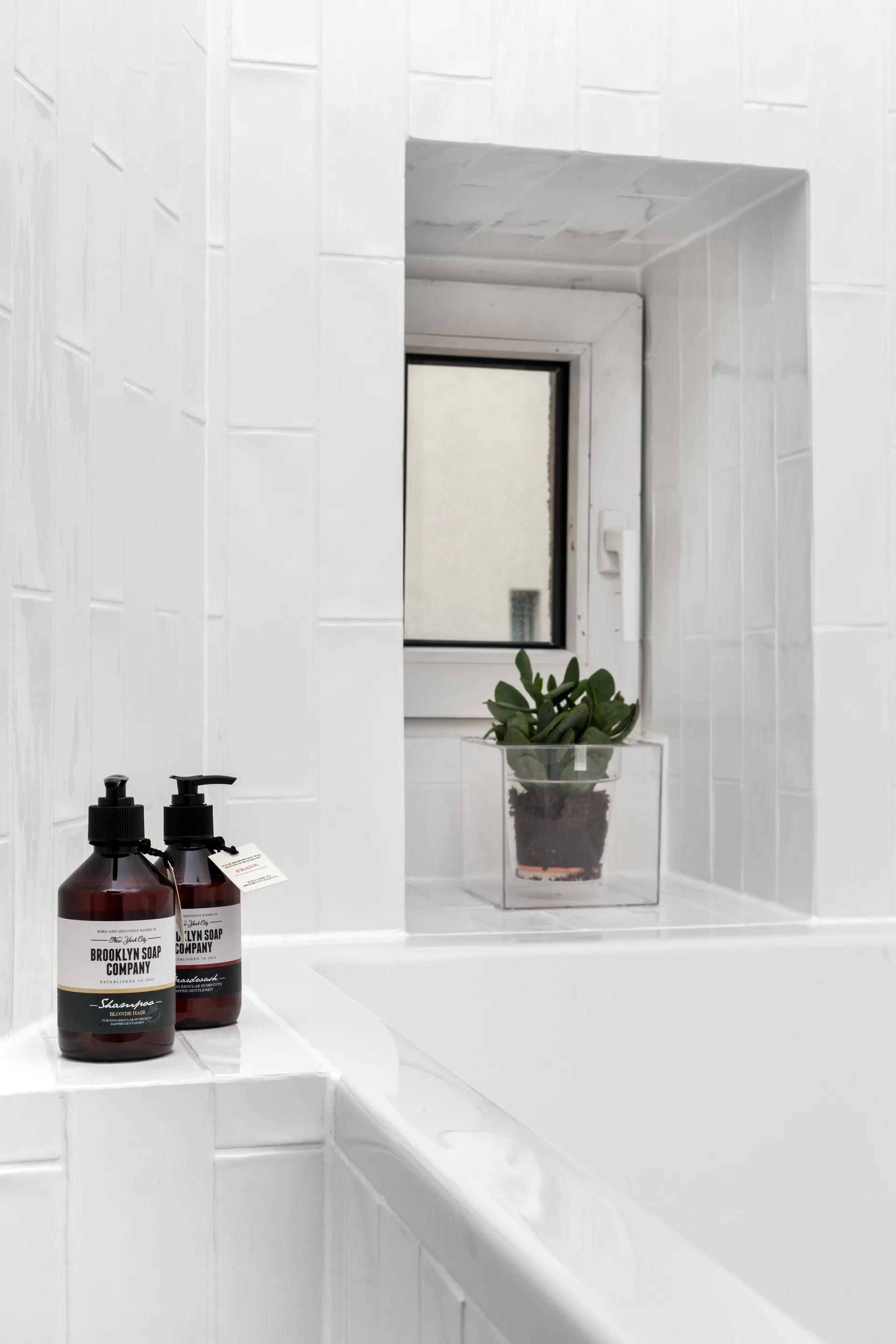 A close-up of vertical white subway tiles in a bathroom, featuring a recessed window niche with a small potted plant and two brown soap bottles.