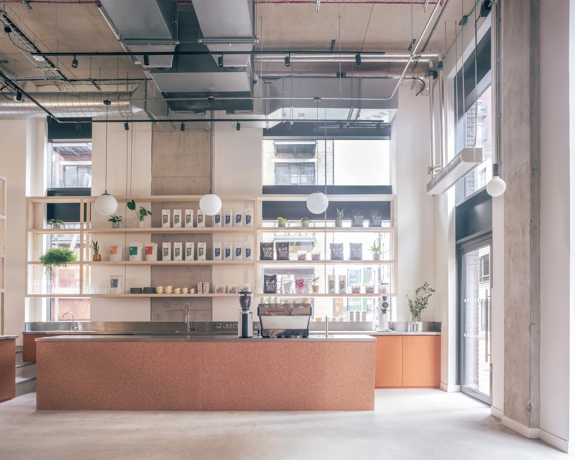 A modern commercial coffee bar featuring an orange terrazzo counter, open light-wood shelving against large windows, and an exposed industrial ceiling.