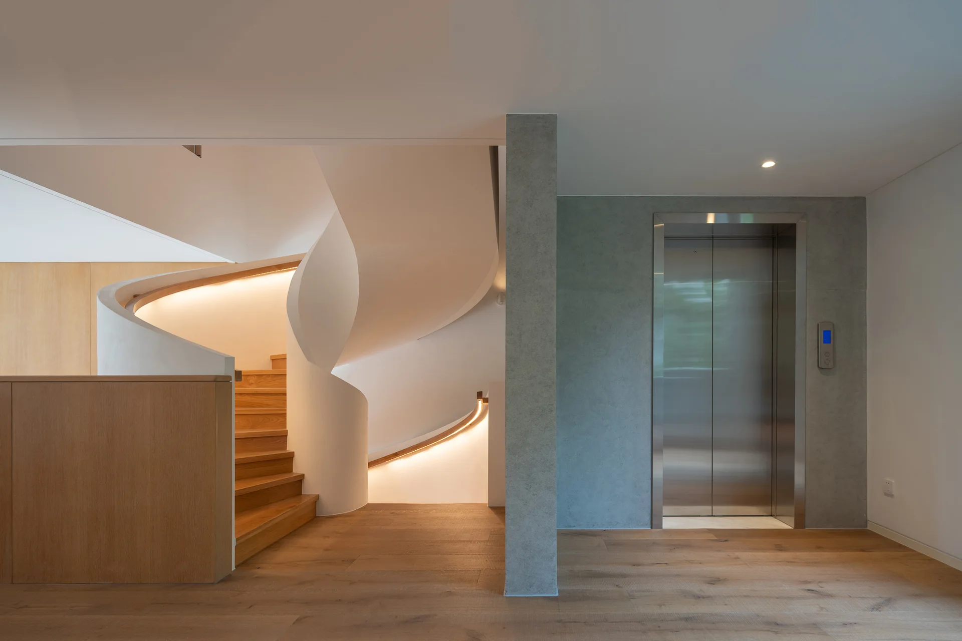 A modern hall features a curved white spiral staircase with glowing handrail lighting and wood treads beside a concrete column and a silver elevator.