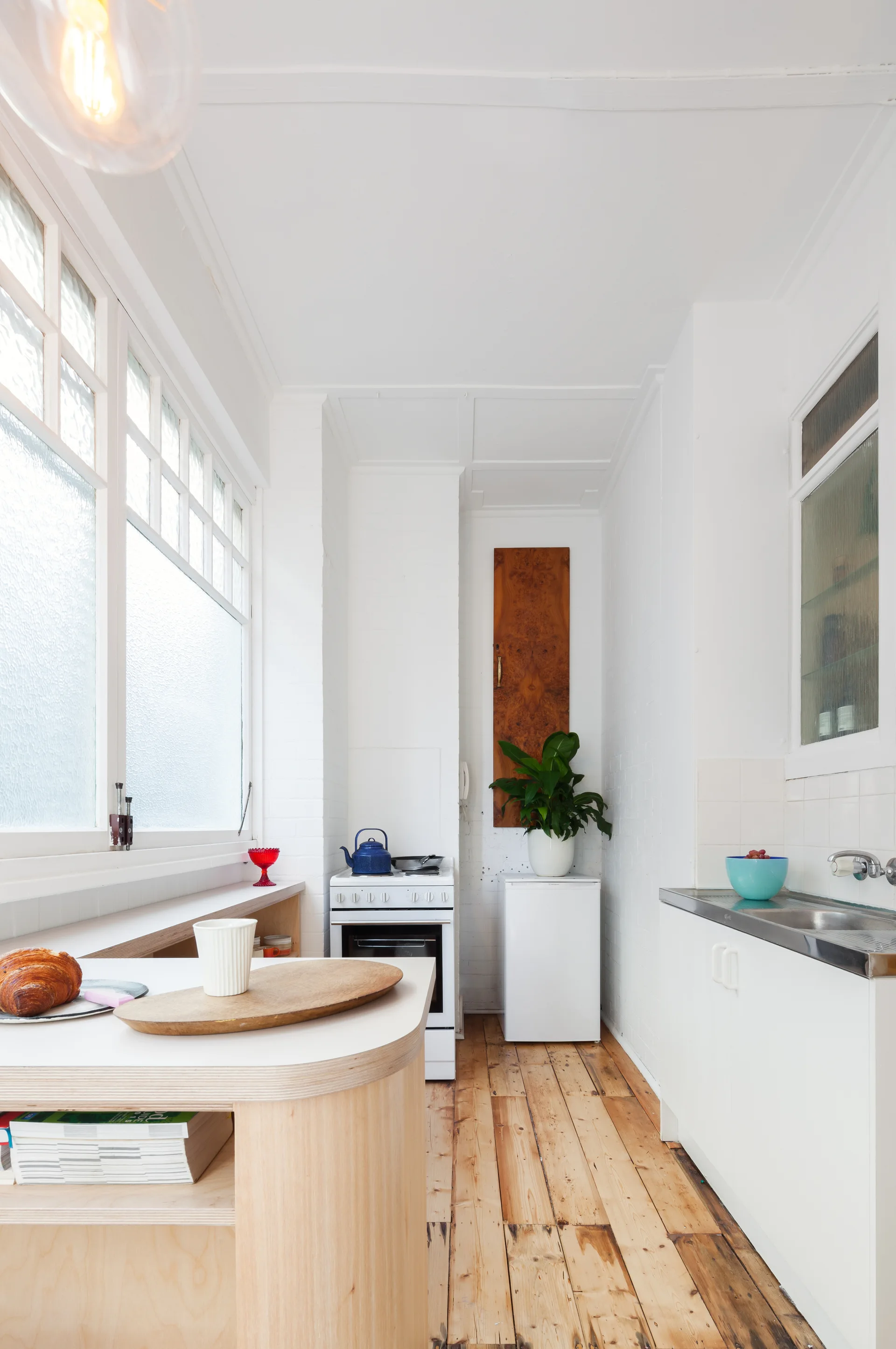 A narrow white kitchen featuring a curved light wood island, exposed wooden floors, a small white stove, and a large frosted glass window.