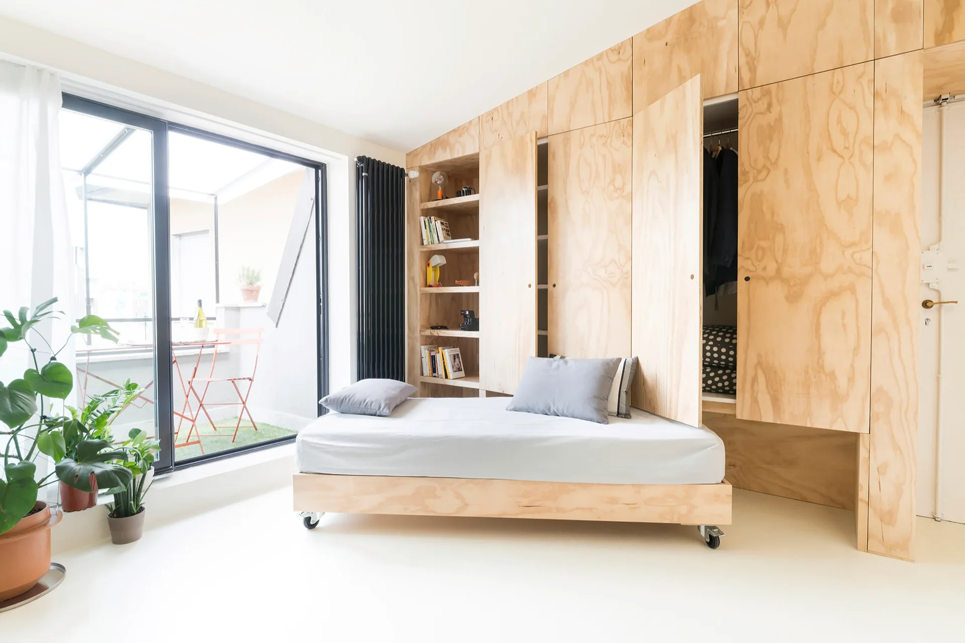 A minimalist white bedroom features a plywood built-in storage wardrobe and a matching low platform bed equipped with black wheels.