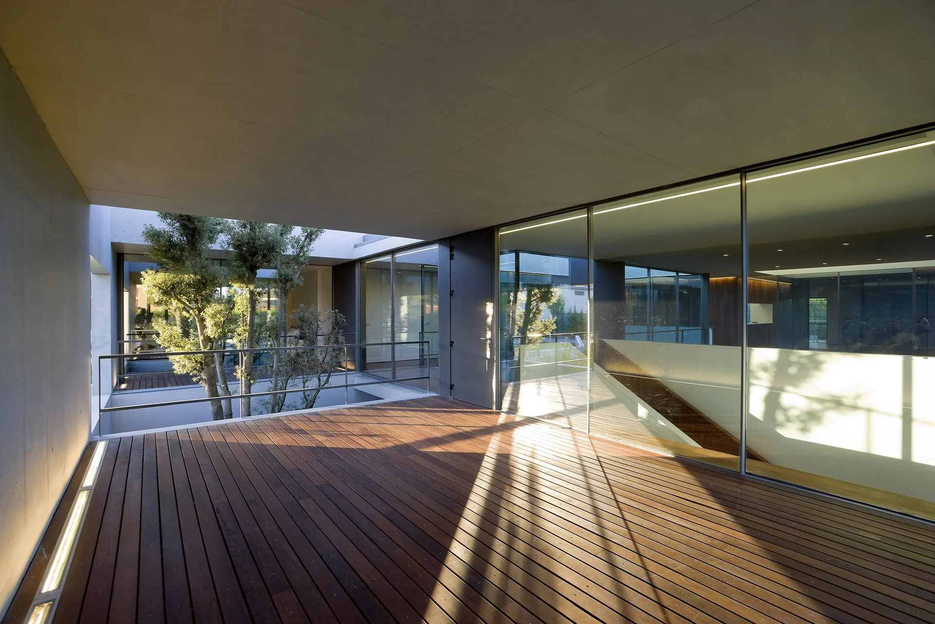 A modern outdoor terrace features a dark wood plank deck, a concrete overhang ceiling, and floor-to-ceiling glass walls looking into the interior.