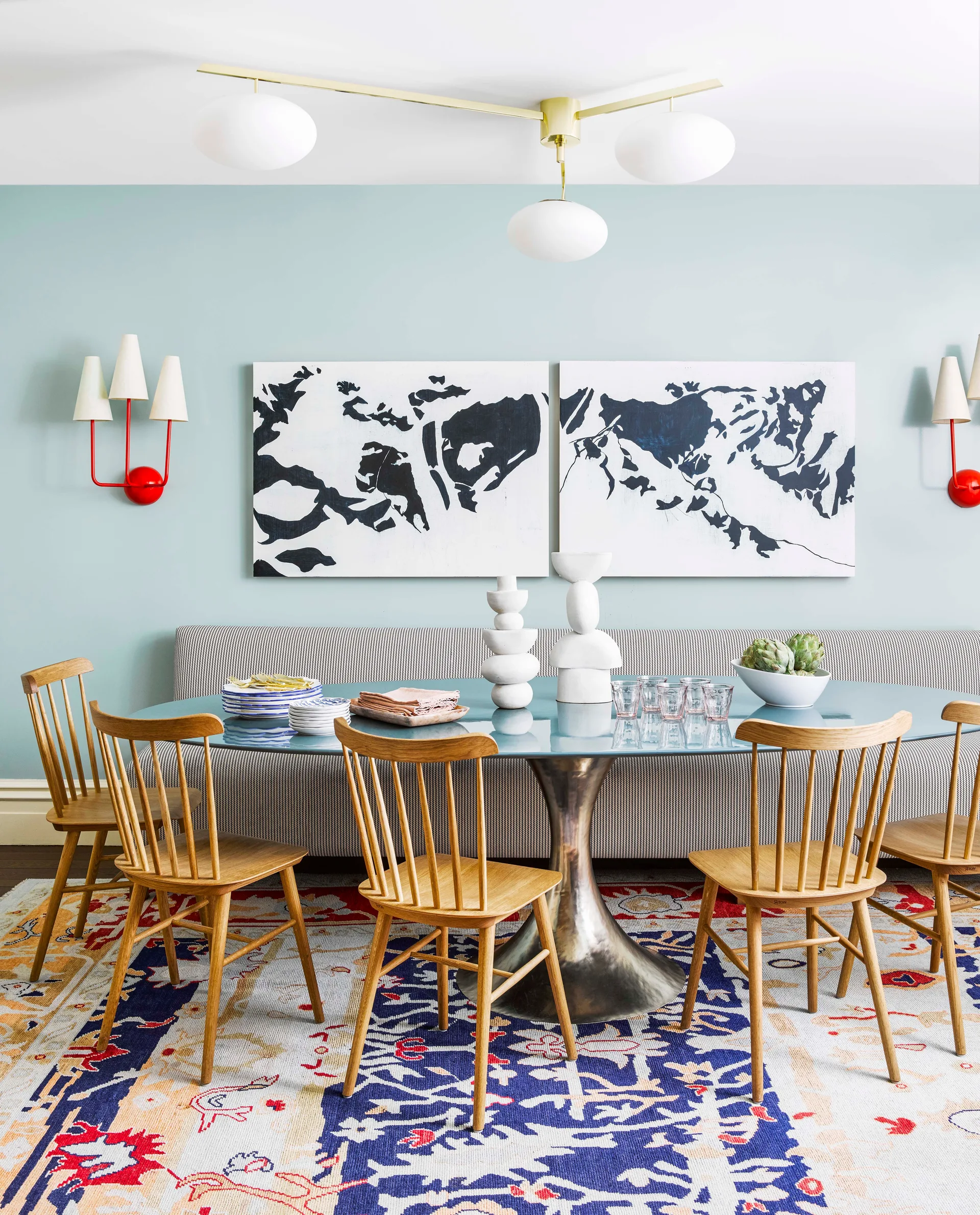 A dining room features a glass-topped pedestal table surrounded by wooden spindle chairs, a striped banquette, and a colorful patterned area rug.