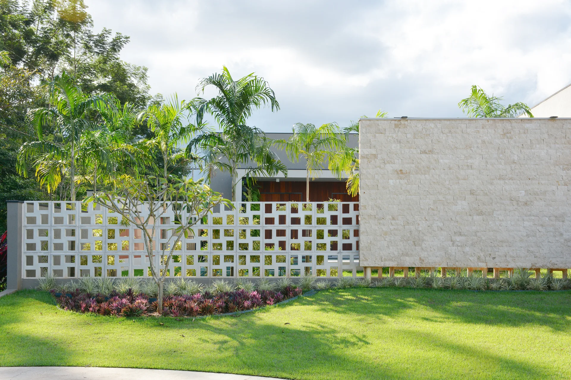 A green lawn sits before a white geometric breeze block screen and a textured beige stone wall under a cloudy sky.
