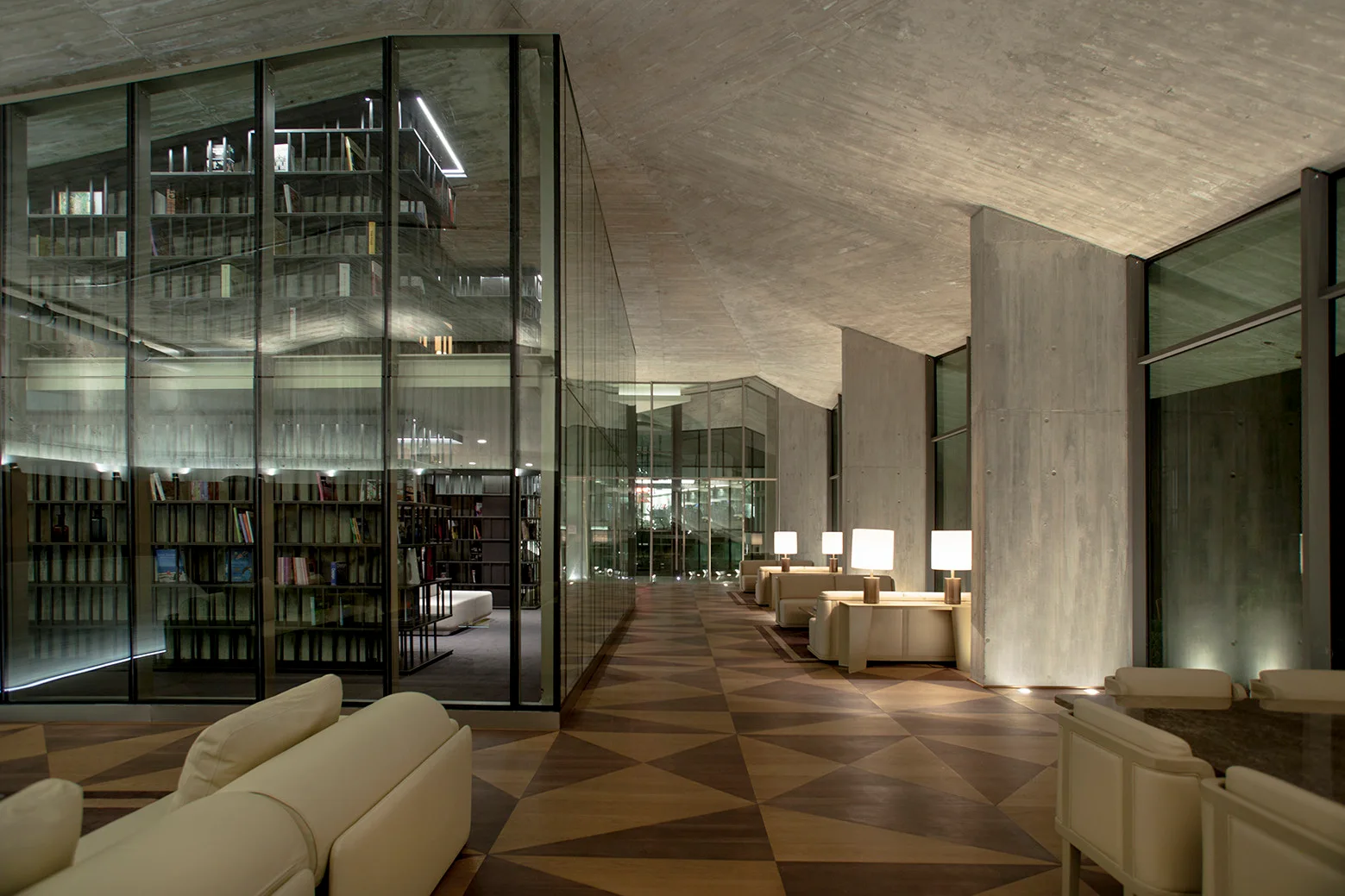 A spacious modern lounge featuring raw concrete walls, a glass-enclosed central library, and a brown geometric patterned wood floor.