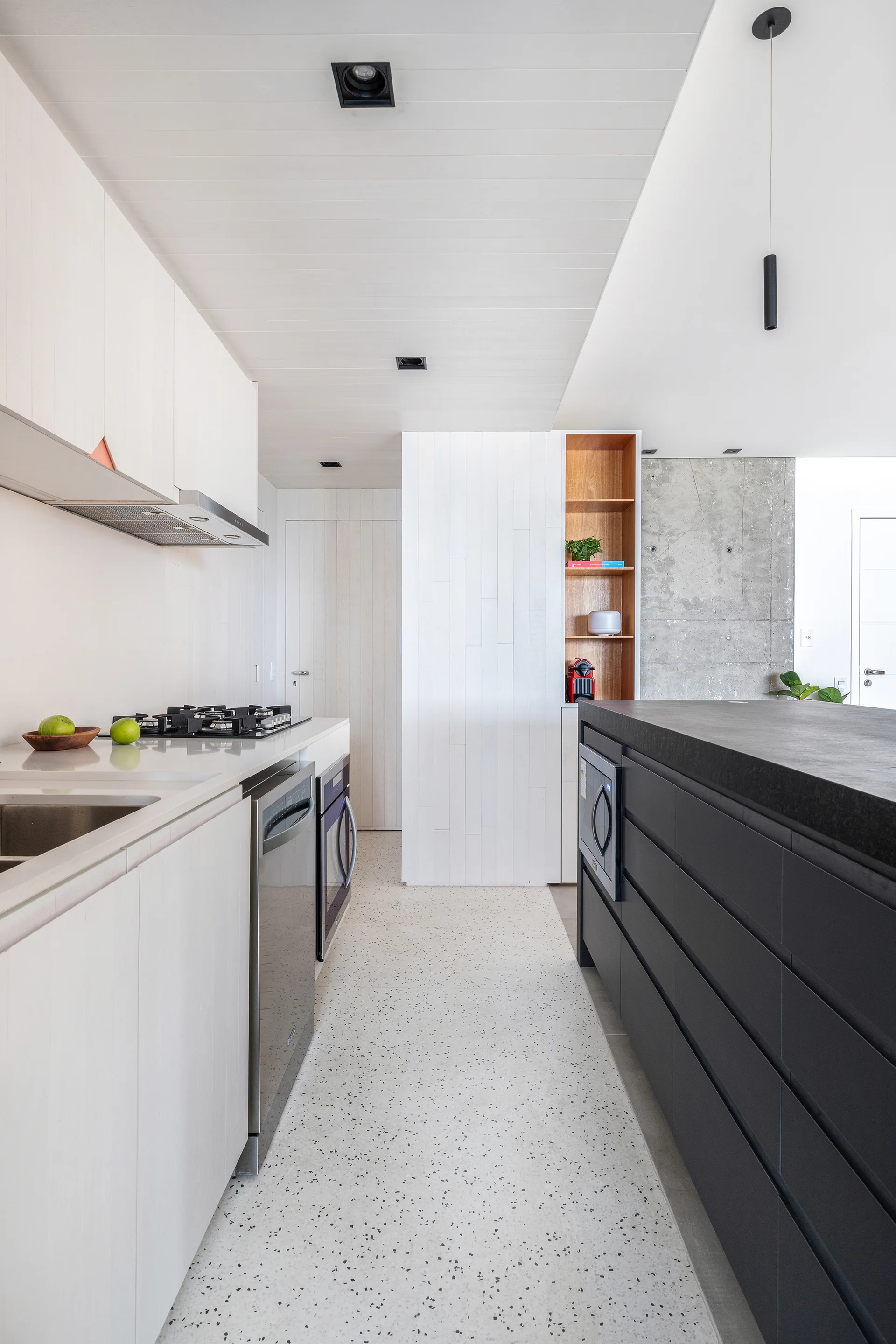 A minimalist kitchen featuring white wood-grain cabinets, a contrasting dark charcoal island, a speckled terrazzo floor, and an exposed concrete wall.