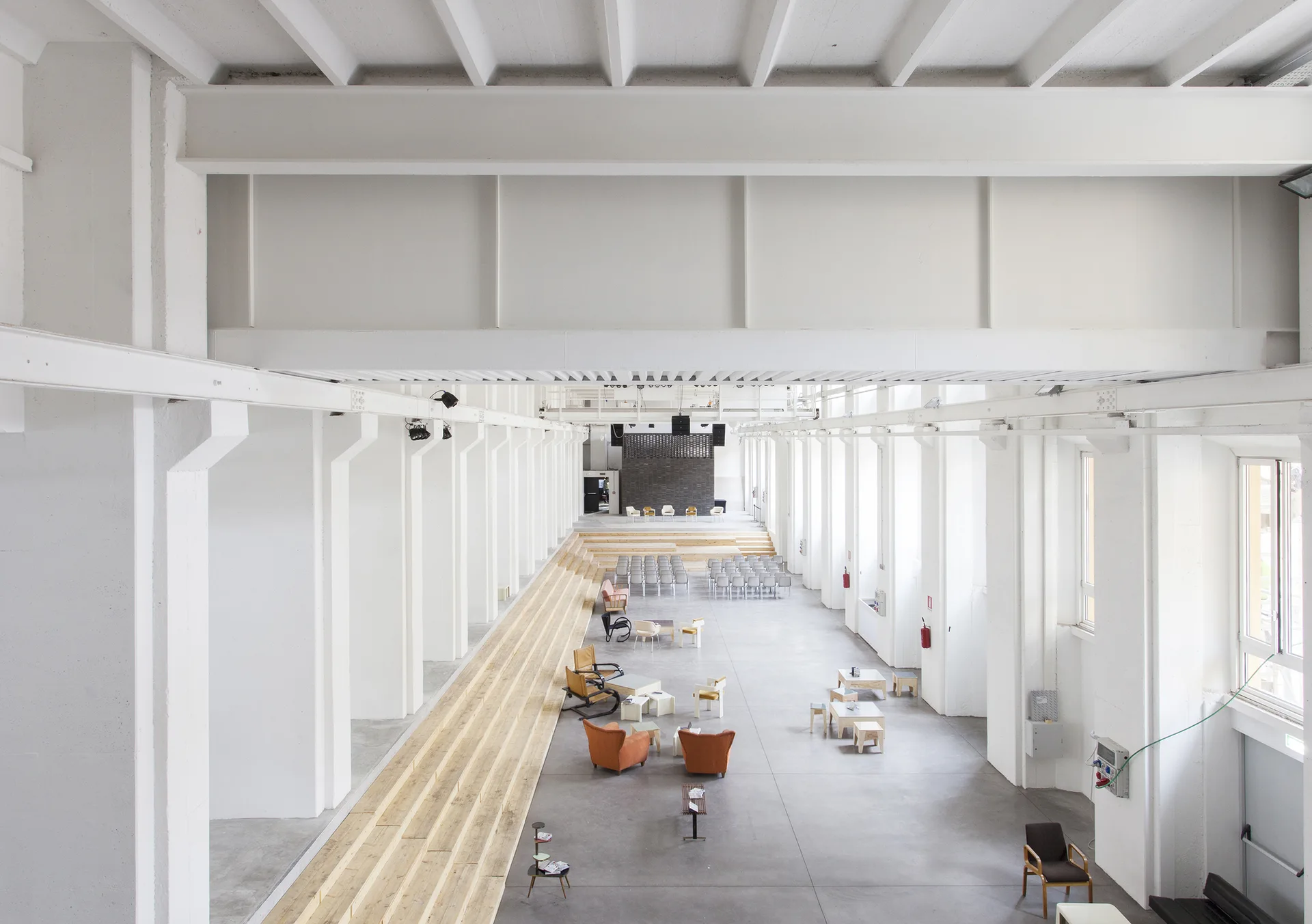 A large industrial open-plan space features structural white beams, columns, a tiered wood platform, and seating groupings on a grey concrete floor.