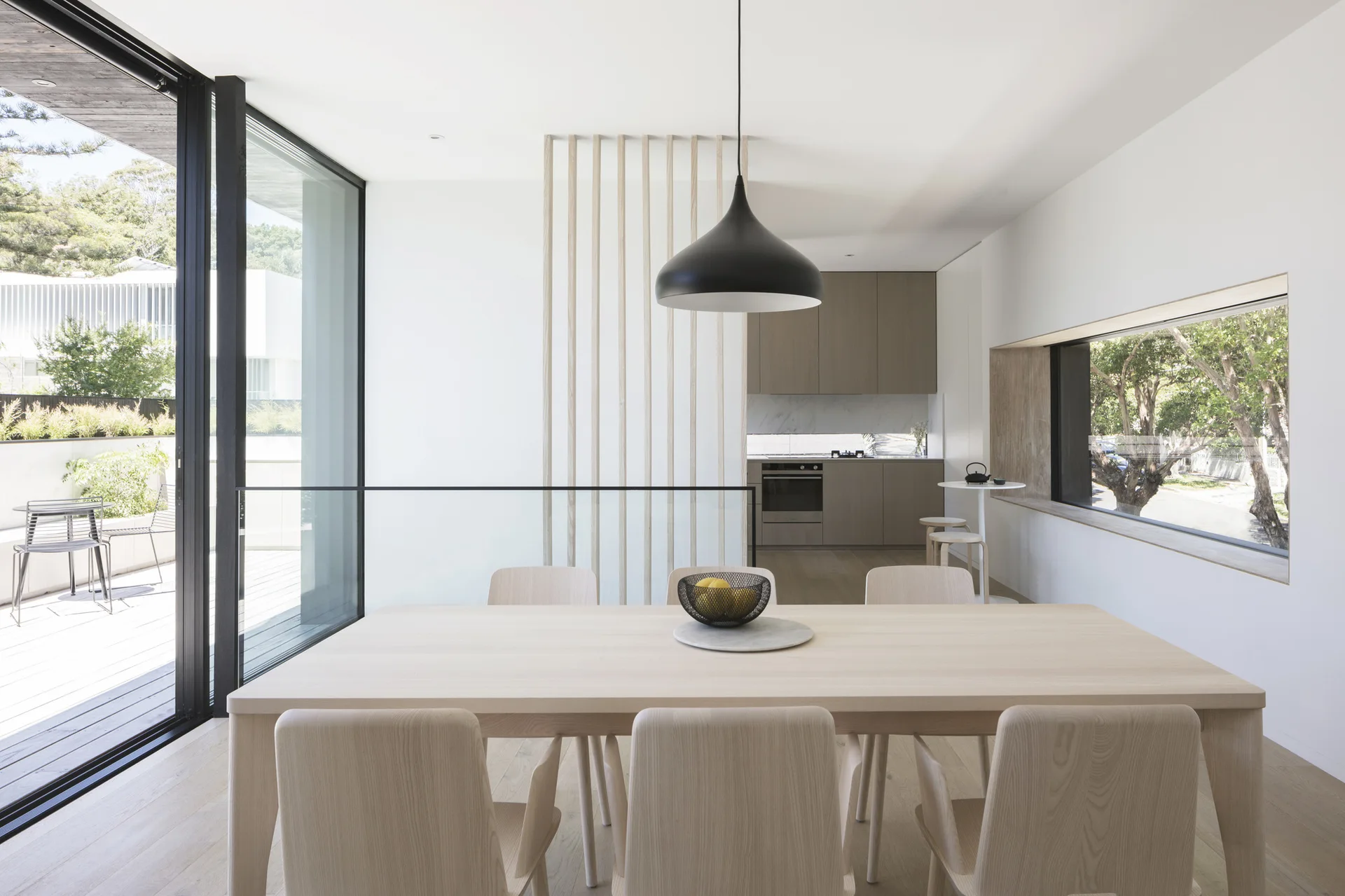 A light wood dining table and matching chairs sit under a black pendant lamp, with a slatted room divider and kitchen in the background.