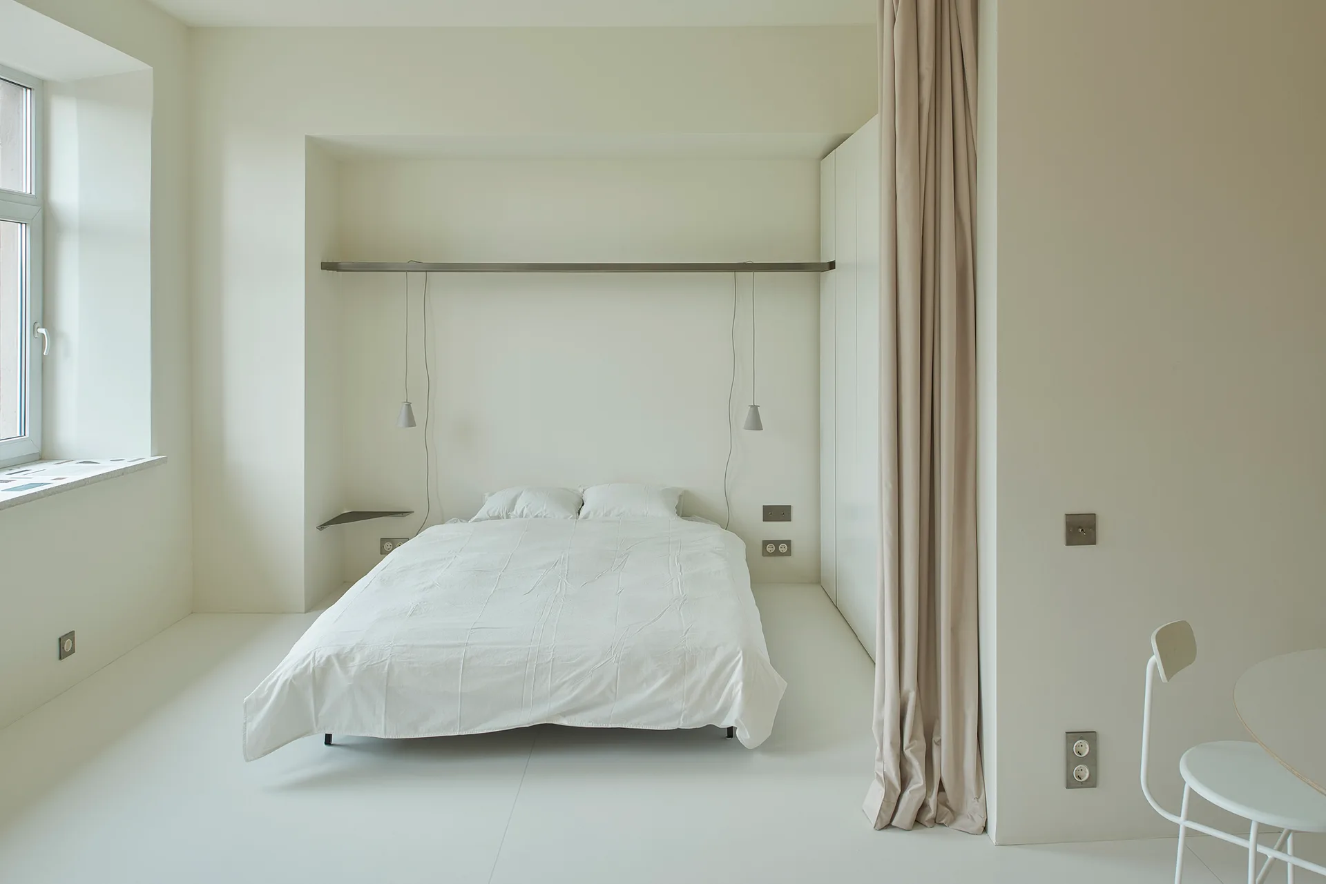 A minimalist all-white bedroom features a recessed bed niche, pendant lights hanging from a long metal shelf, and a beige room-dividing curtain.