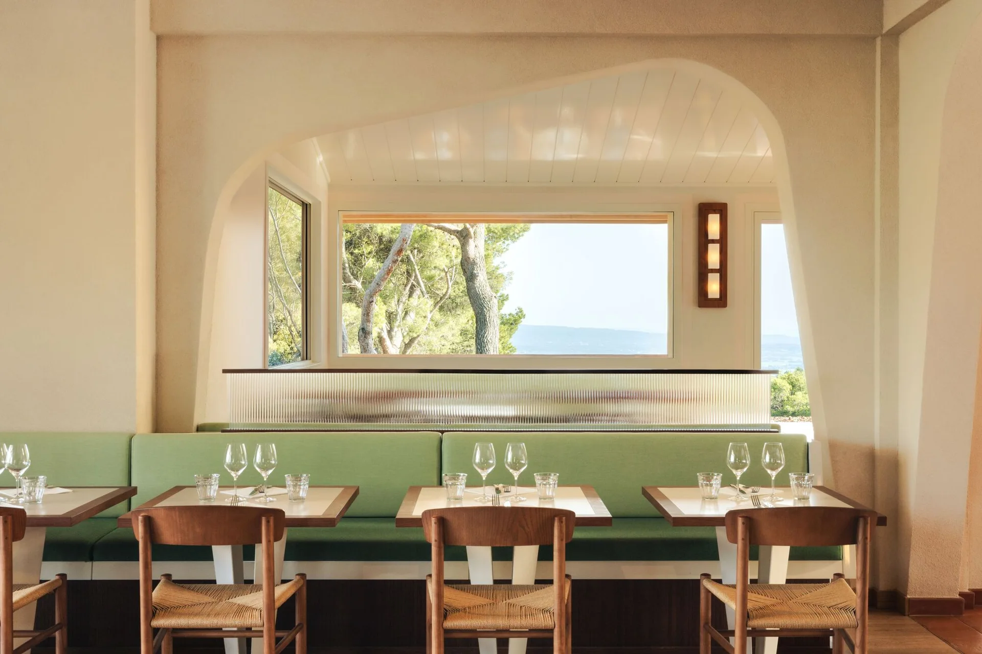A restaurant dining area features sage green banquette seating, wood chairs with woven seats, and a large window framed by a curved plaster archway.