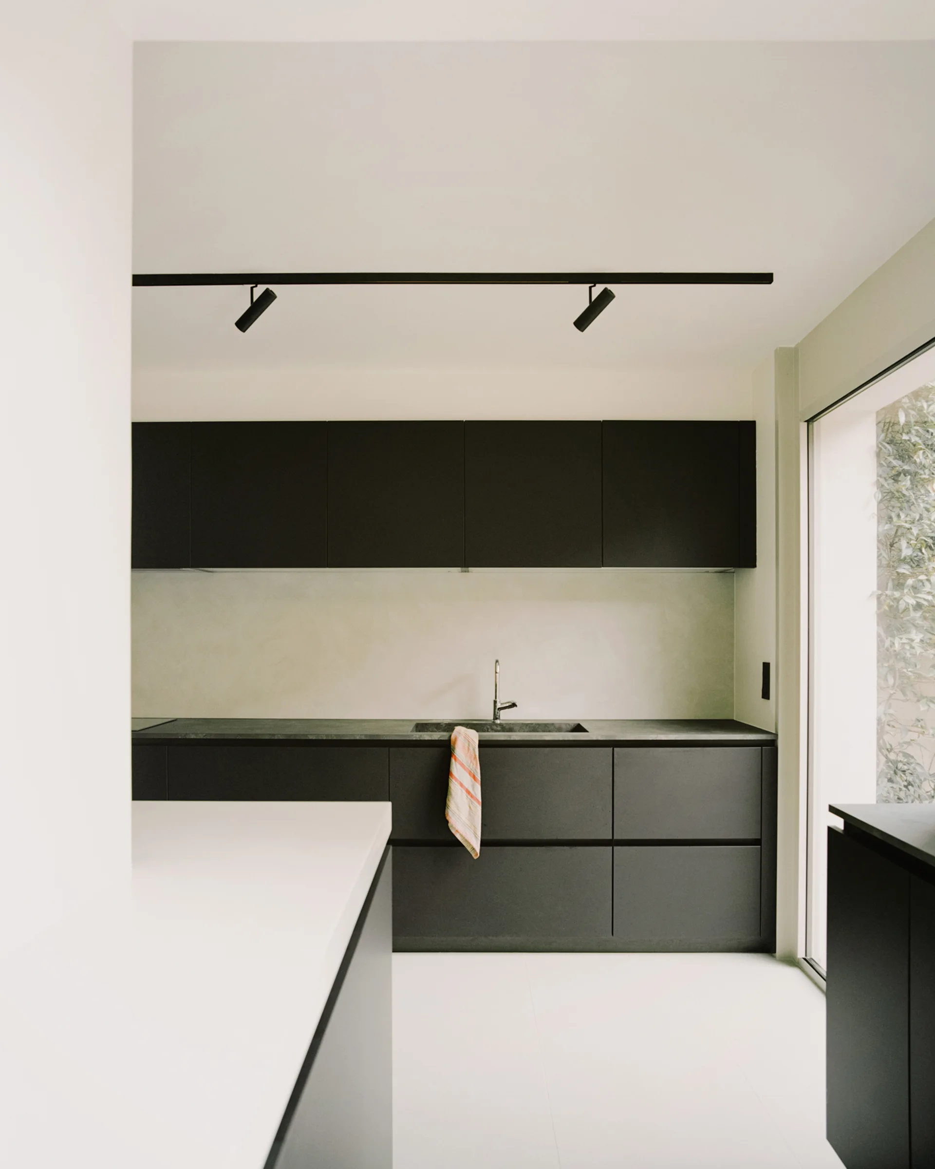 A minimalist kitchen featuring matte black handleless cabinets, a dark integrated countertop with a sink, white tiled floors, and black ceiling track lights.