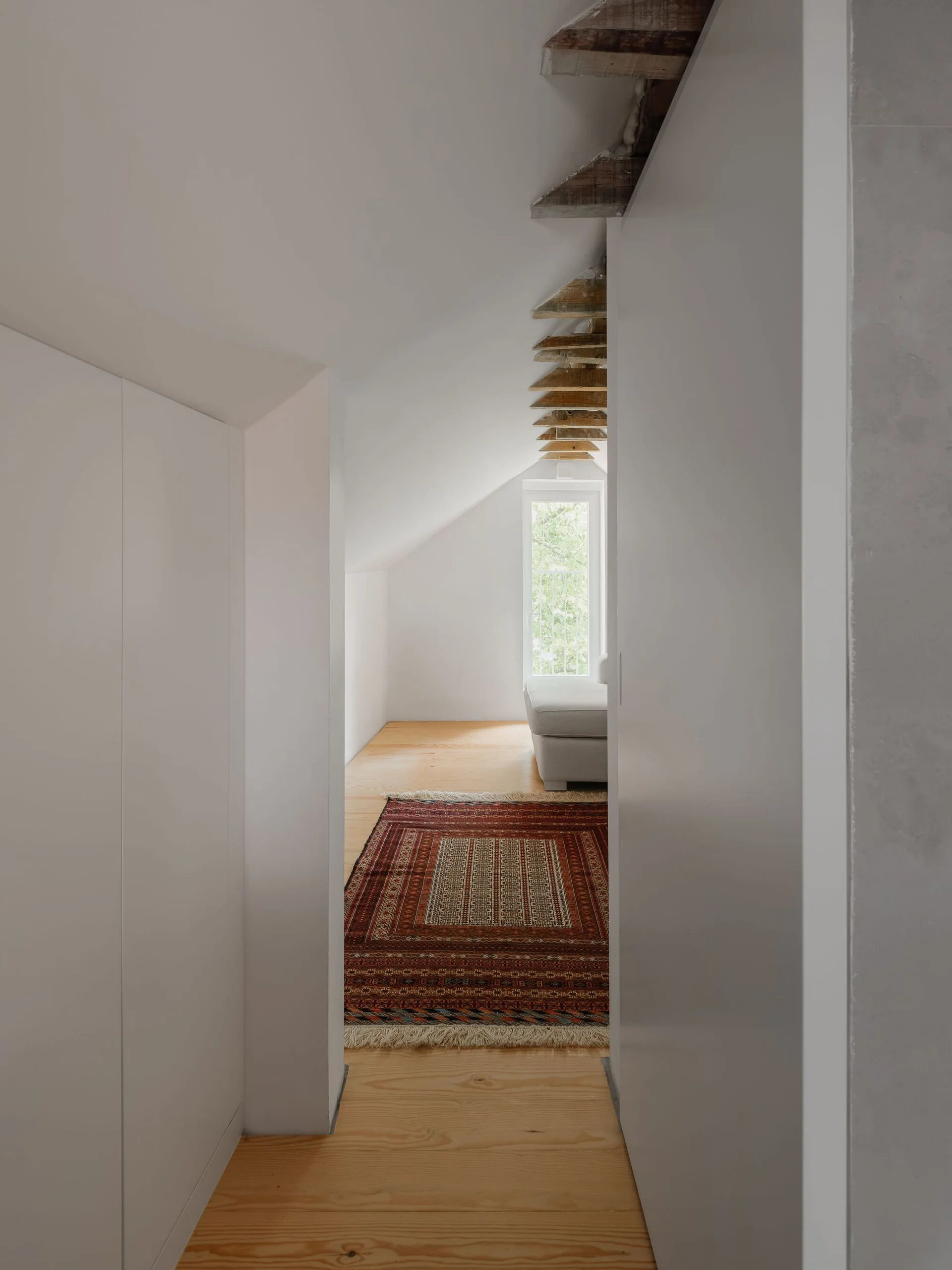 A white attic room with light wood flooring, a red patterned rug, exposed wooden rafters, and a grey sofa near a window.