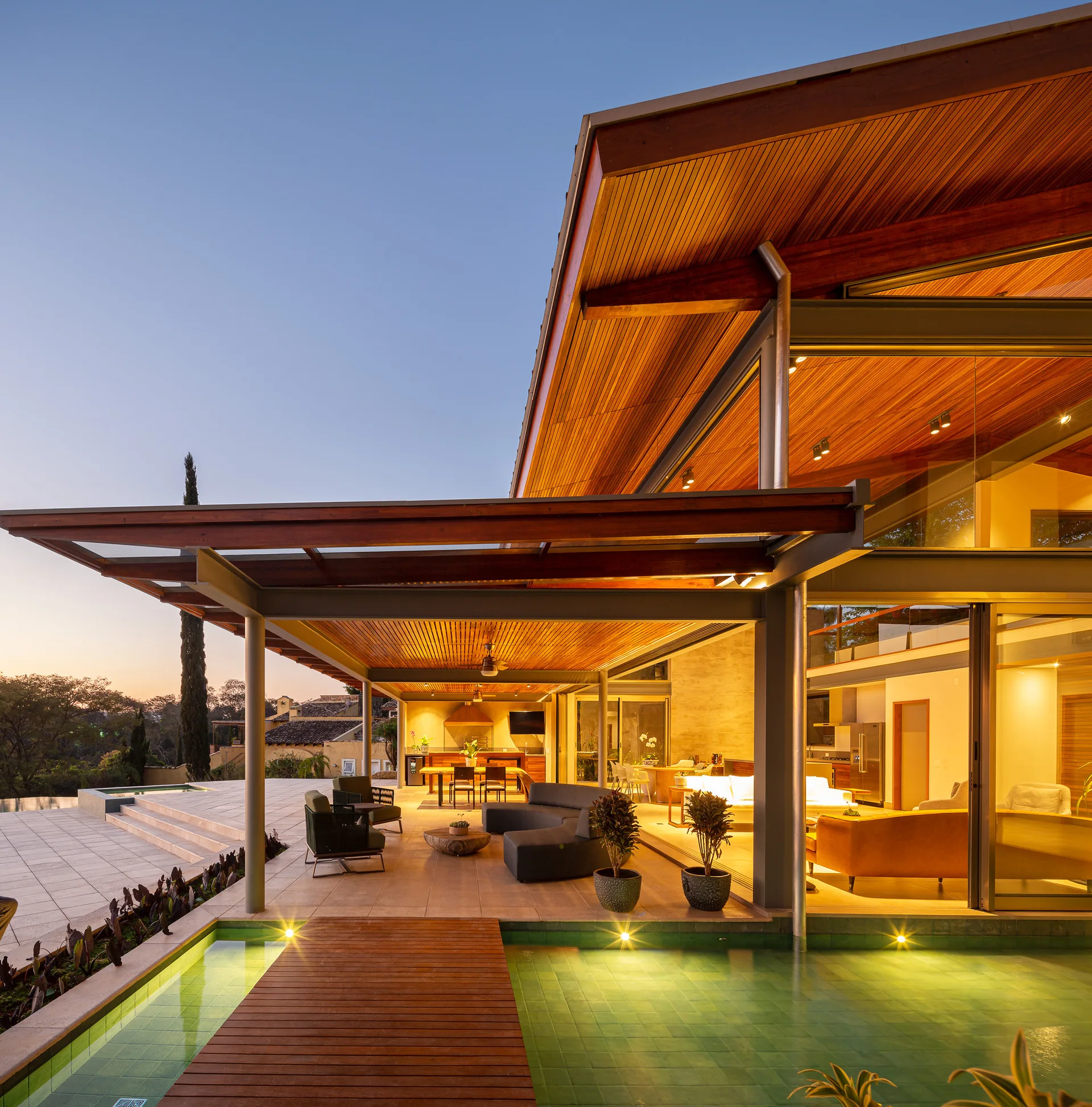 A modern covered patio features warm slatted wood ceilings, a curved outdoor sofa, and a wooden bridge crossing a green-tiled swimming pool.