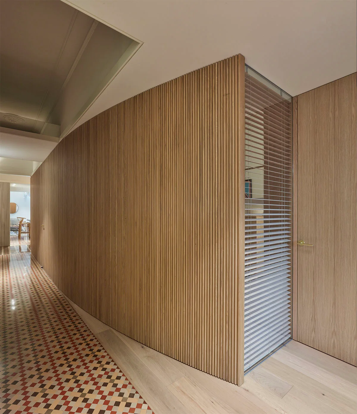 A minimalist hallway features a curved vertical wood slat wall, a flush wood door, and a geometric tiled floor transitioning to light wood planks.